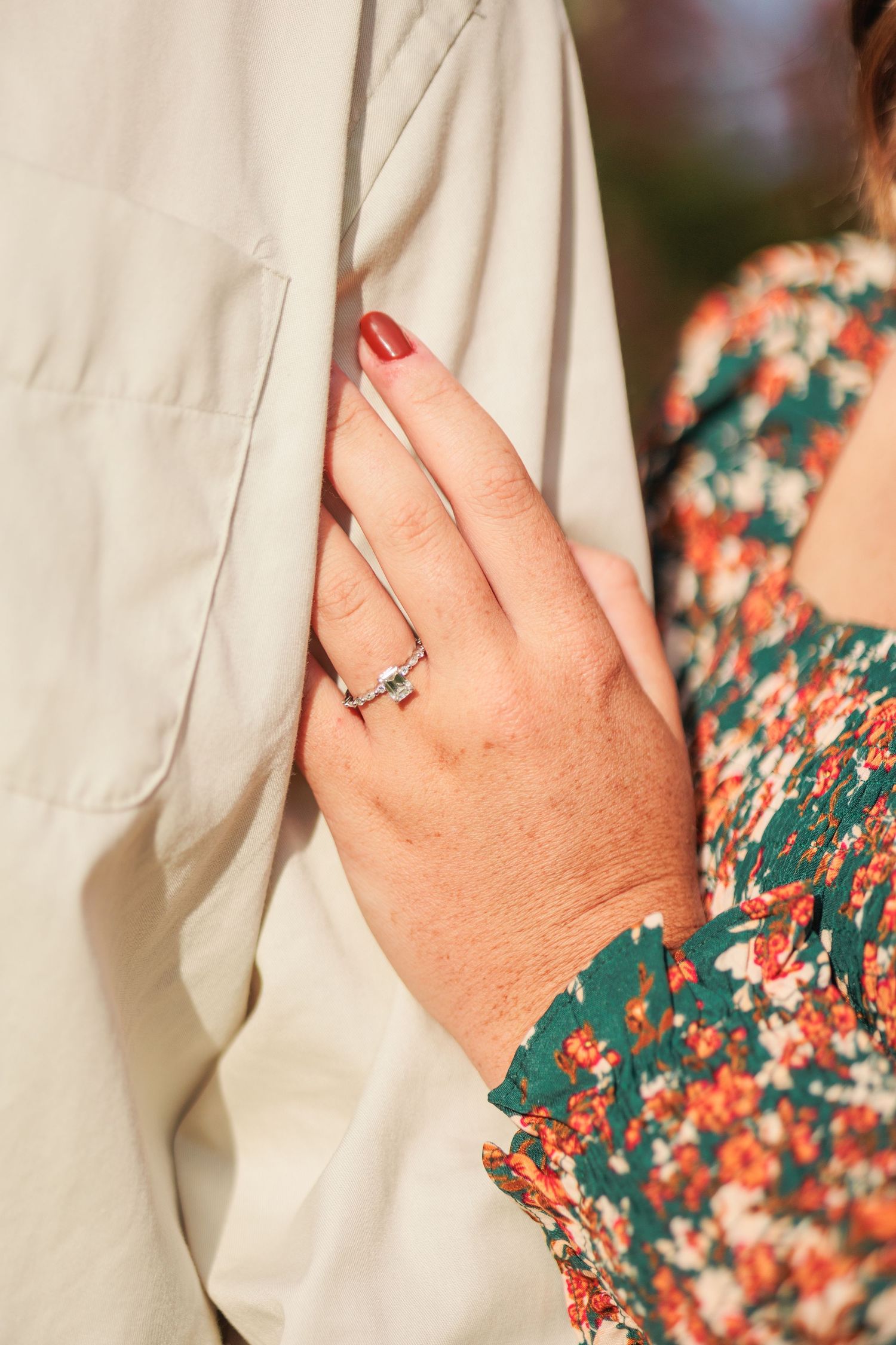 Close-up of womans hand with diamond engagement ring and red nails resting on mans light jacket outdoors.