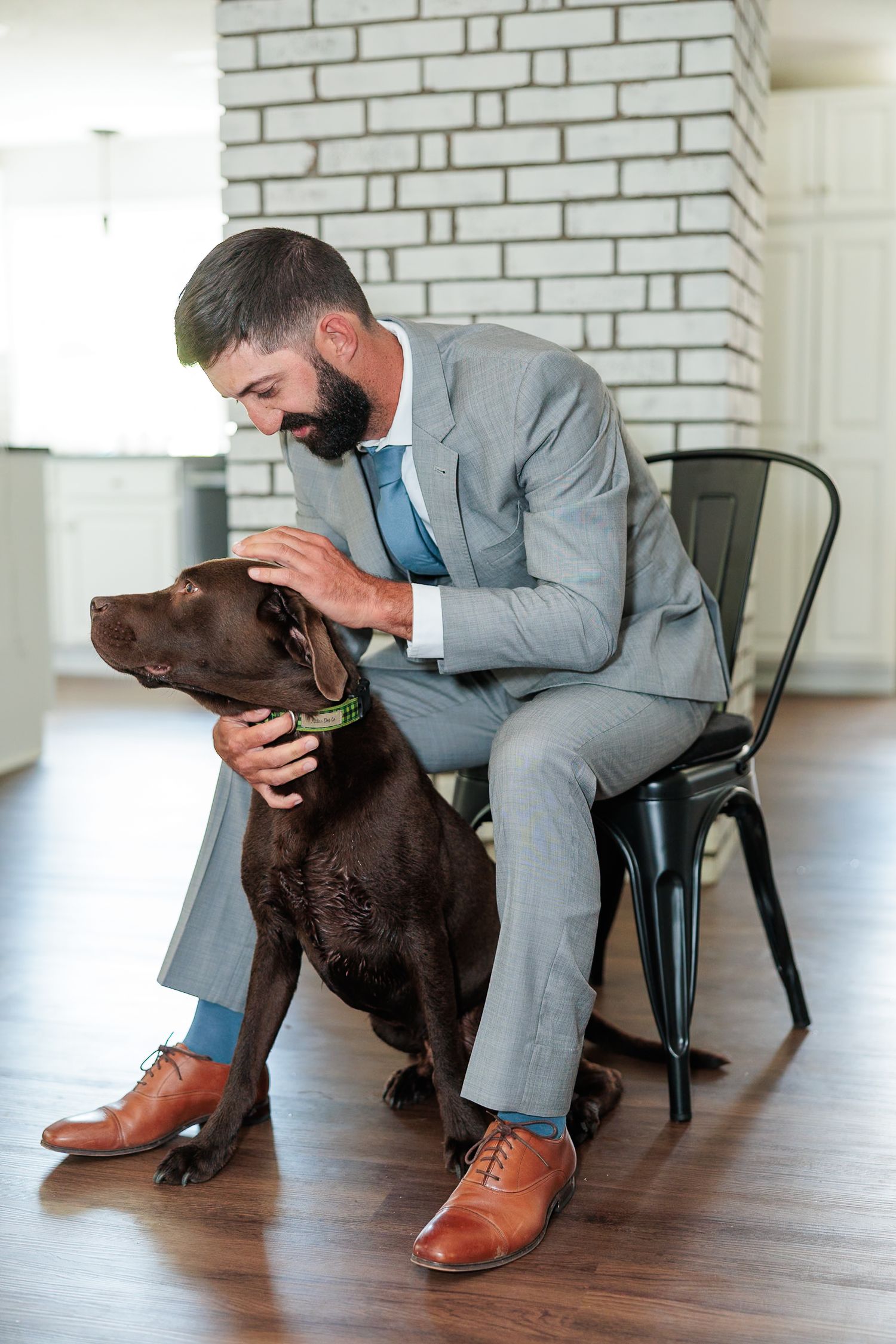 Dapper groom in gray suit and blue tie sitting on chair petting chocolate Labrador dog indoors.