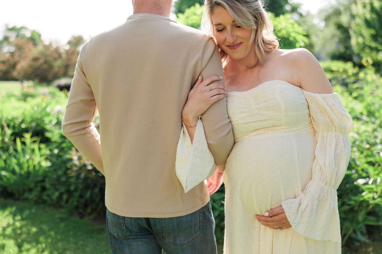 Pregnant woman in elegant white off-shoulder dress standing with partner in a lush green garden.