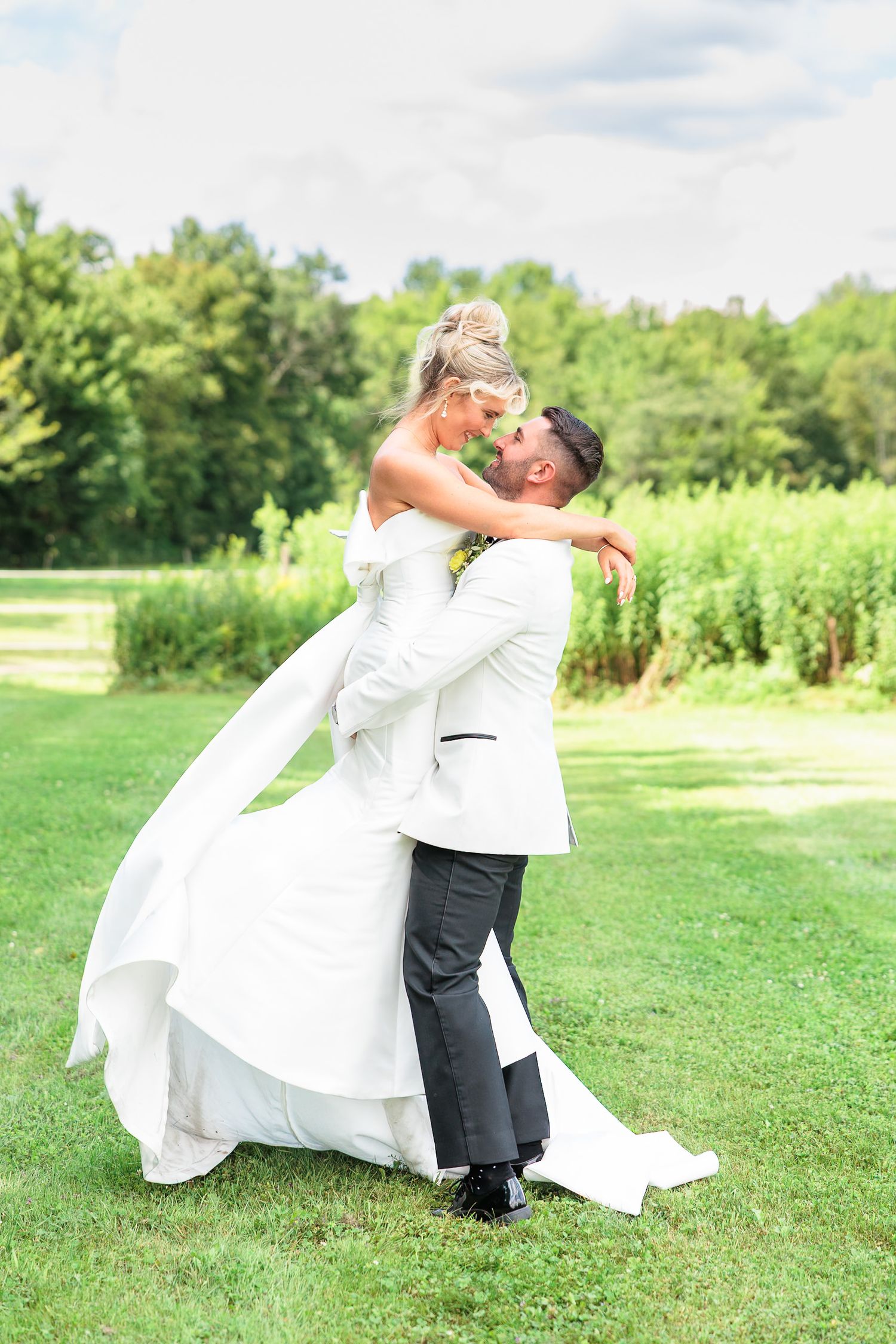 Groom in white jacket lifts bride in elegant white gown during joyful outdoor wedding photo on green lawn.