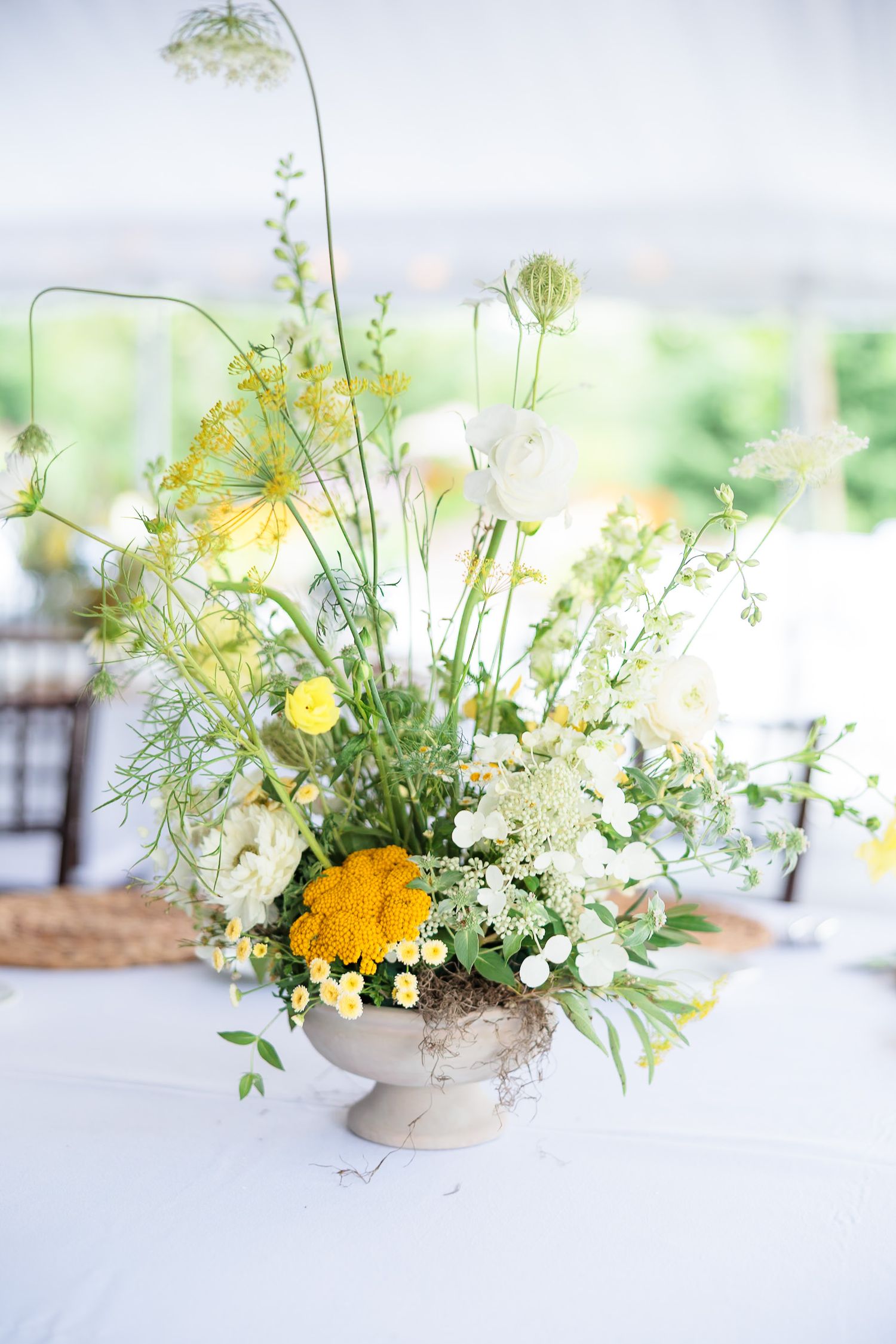 Elegant yellow and white wildflower wedding centerpiece in a stone urn on a white tablecloth.