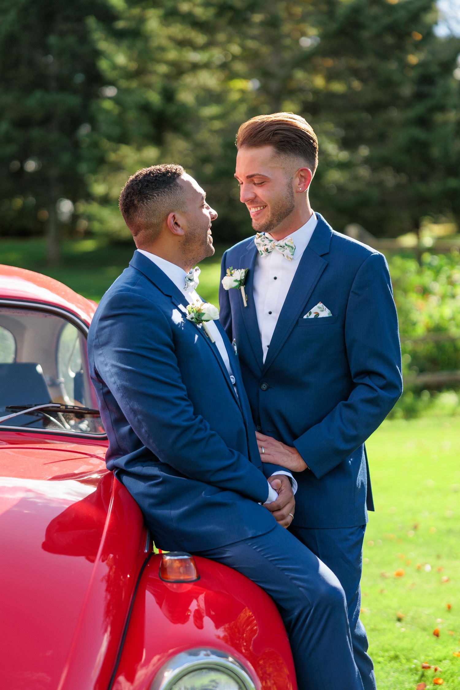 Couple in navy suits share romantic moments beside red vintage Volkswagen Beetle.