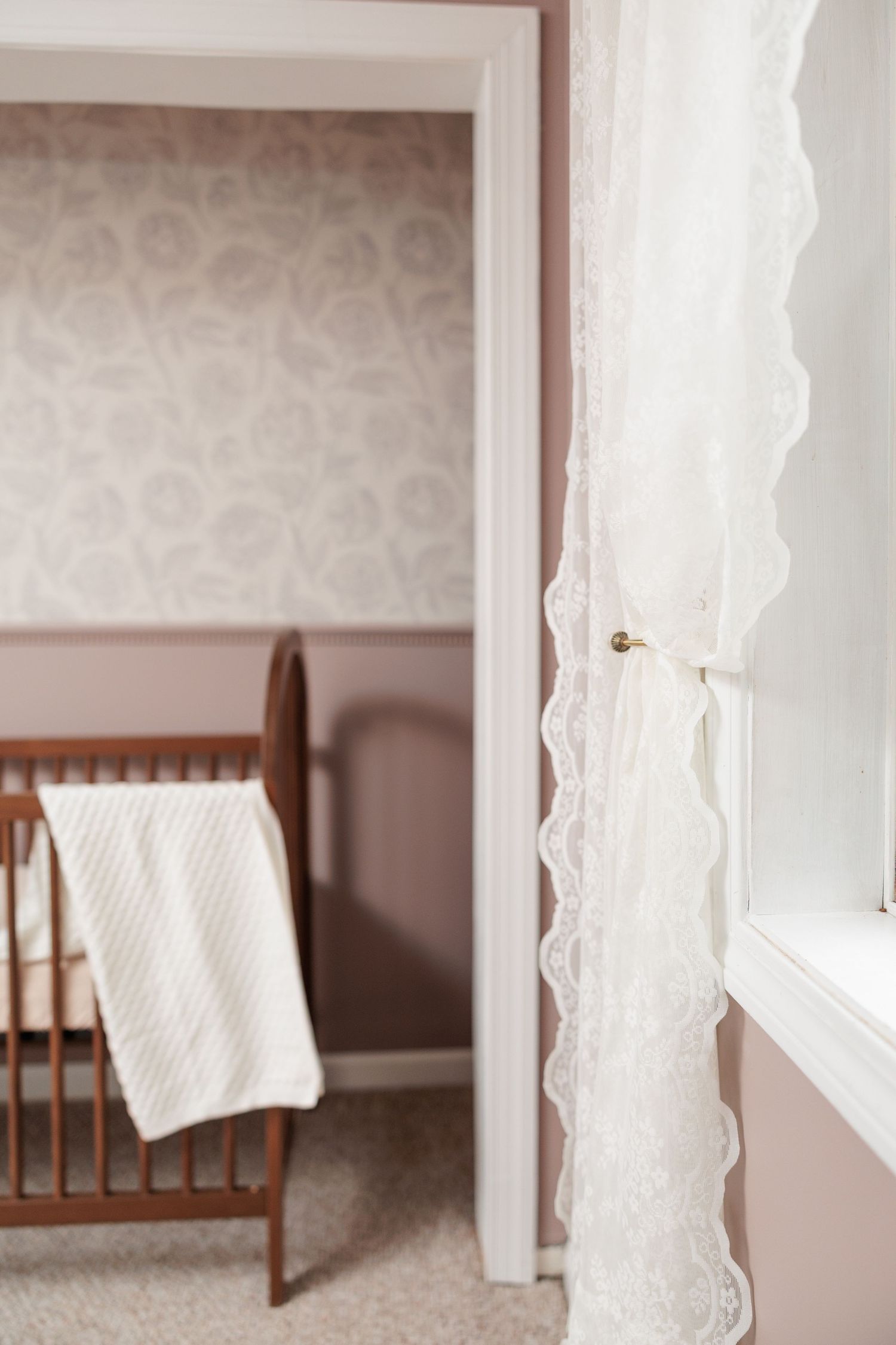 A wooden crib with a white blanket in a nursery room with floral wallpaper and lace curtains.