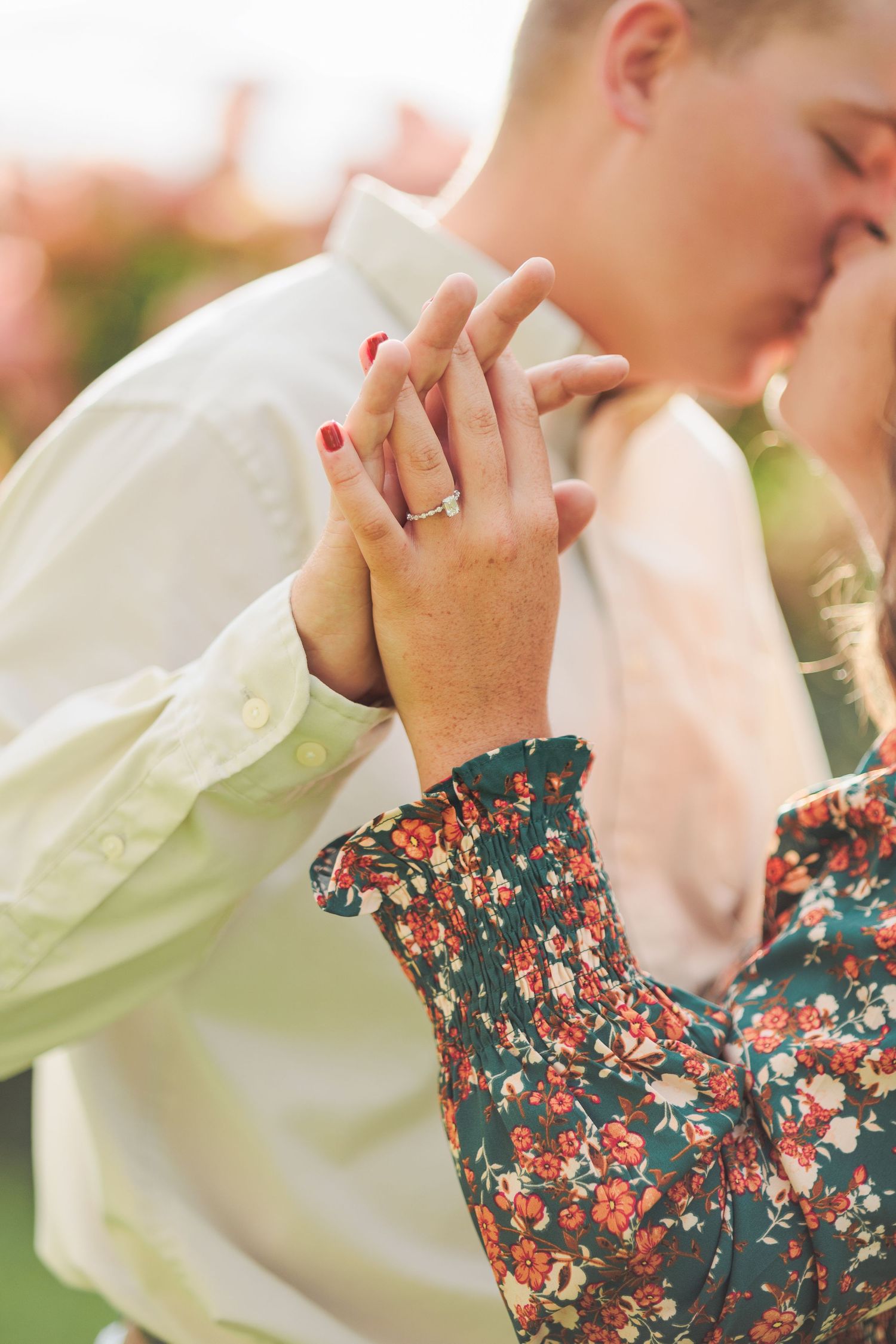 Close-up of engaged couple holding hands showing diamond ring while sharing a kiss outdoors.