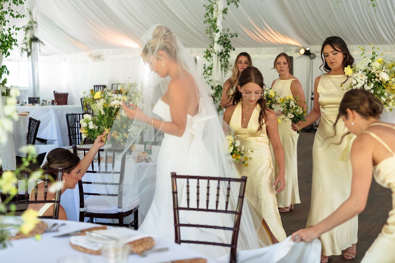 Bride in strapless gown arranges yellow and white bouquets with bridesmaids in yellow dresses in reception tent.