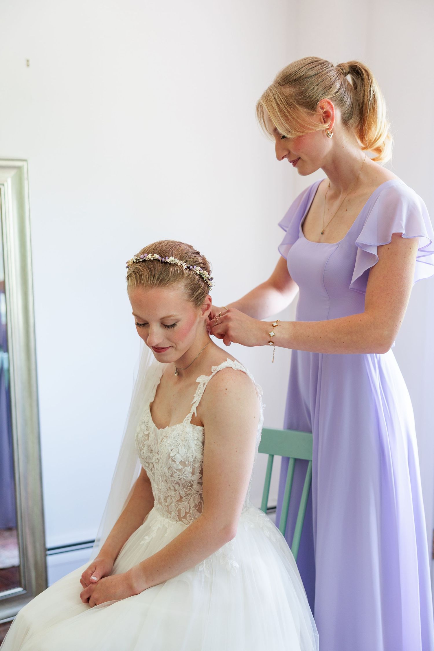 Bridesmaid in lavender dress helps bride with earrings while seated before a mirror getting ready.