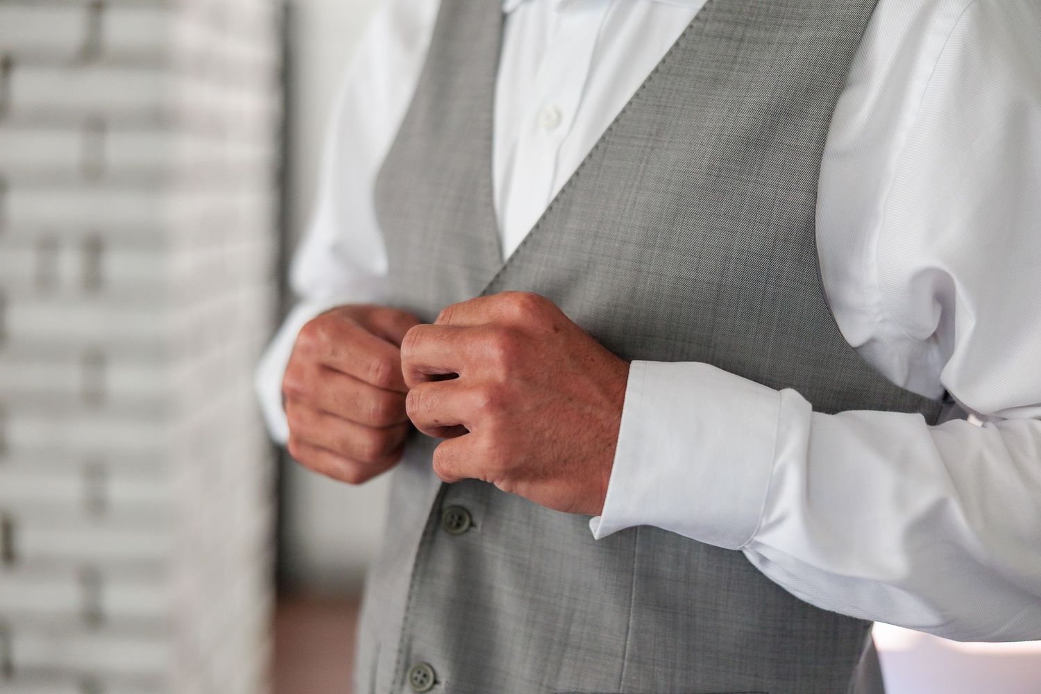 Groom buttoning gray suit vest over white dress shirt against white brick wall background.