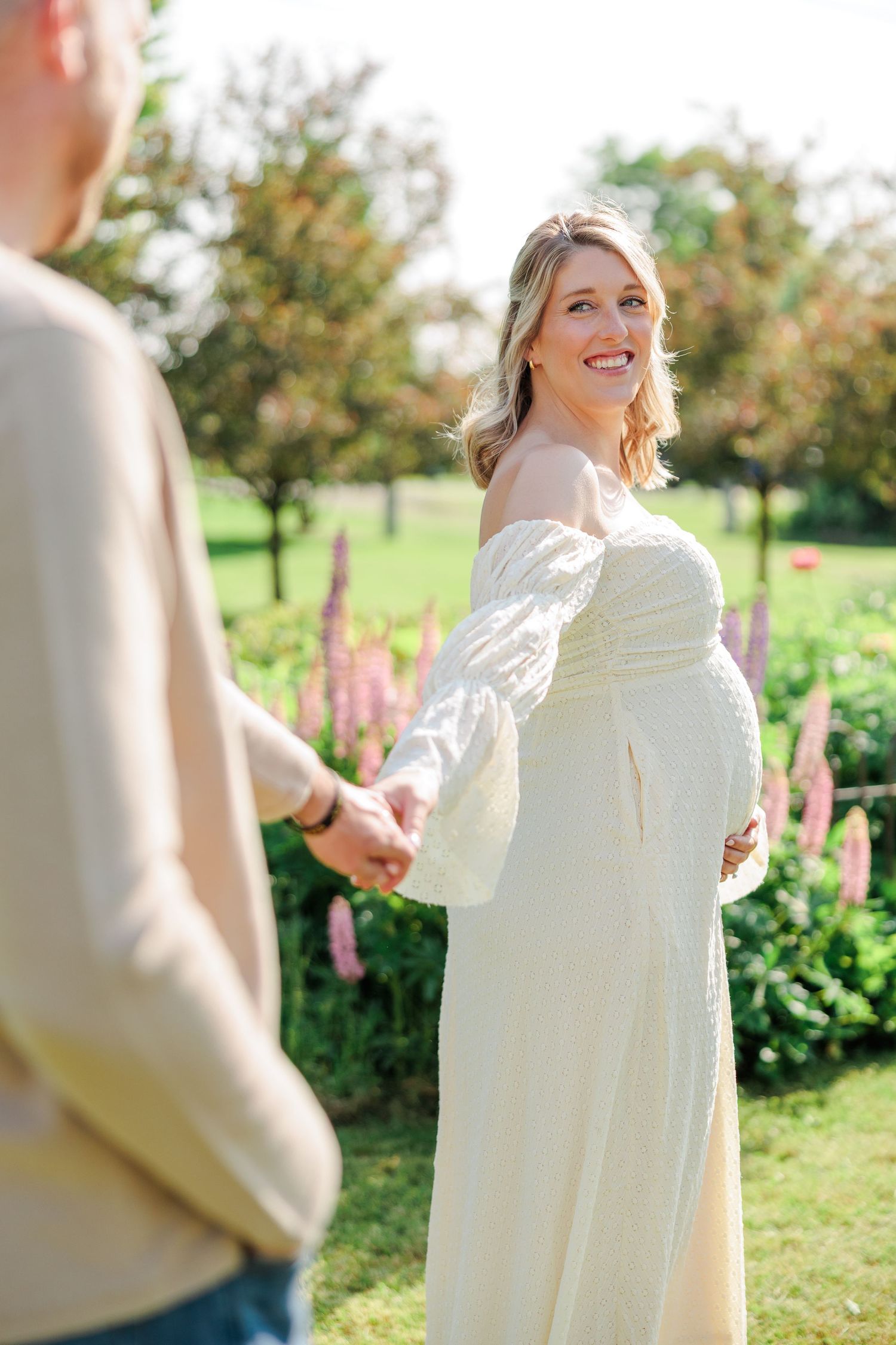 Glowing pregnant woman in white off-shoulder gown smiling while holding partner's hand in sunny garden.