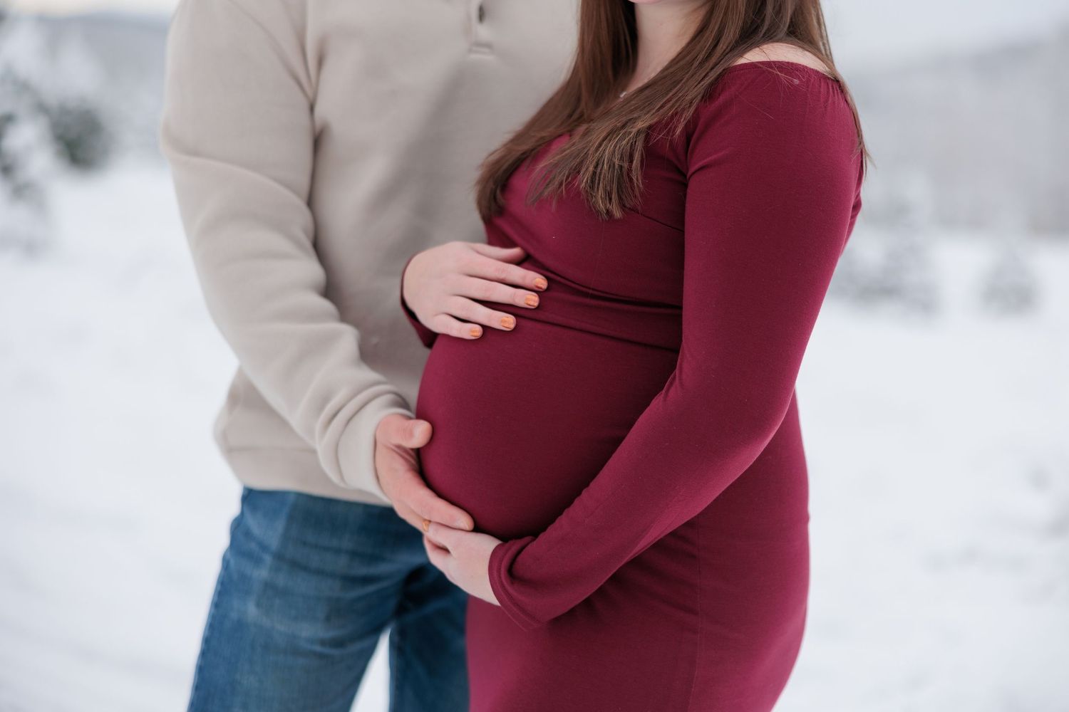 A couple tenderly cradles a pregnant belly in matching burgundy and beige winter outfits outdoors.