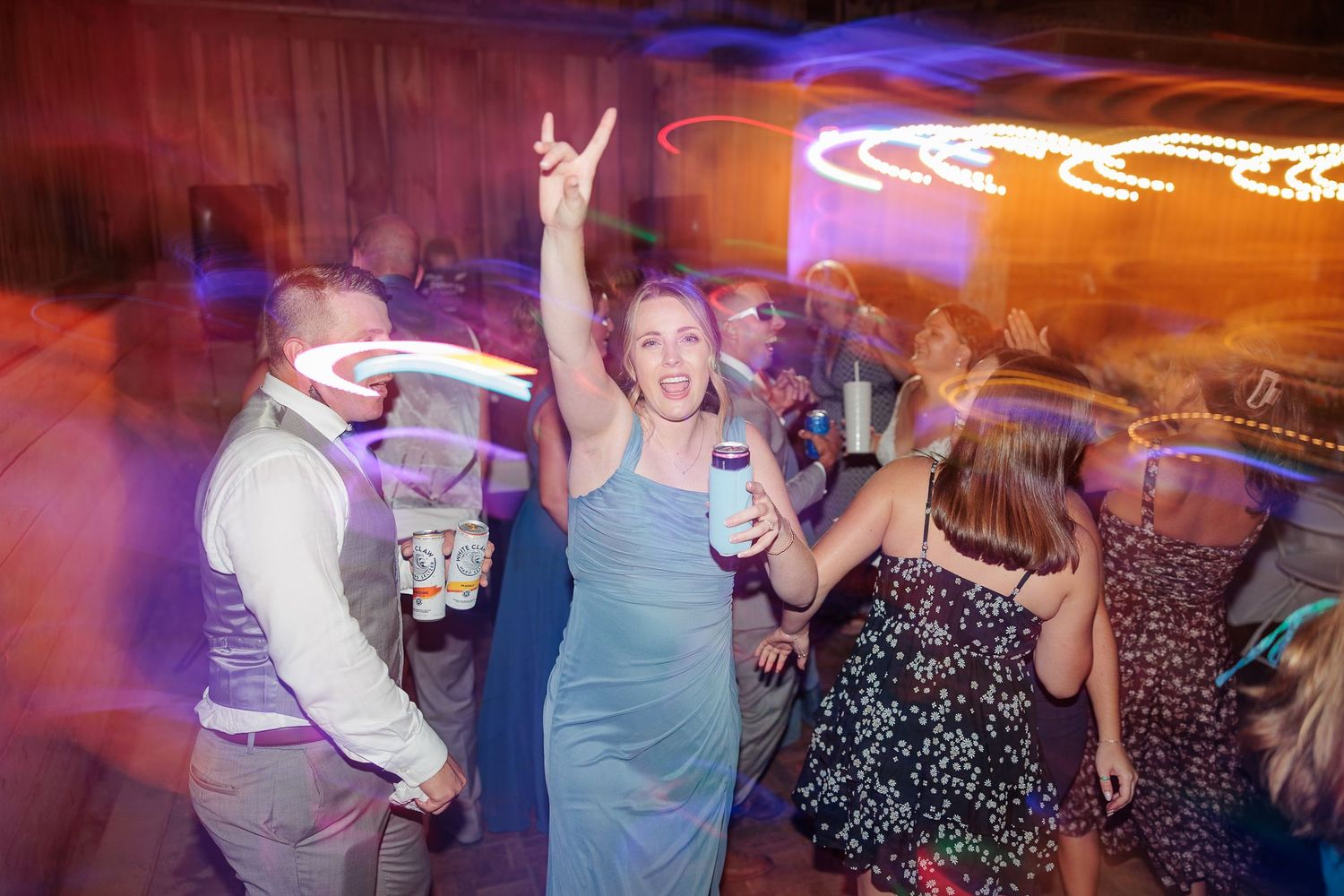 Wedding guest in teal dress celebrates on dance floor with hands up during lively reception party.