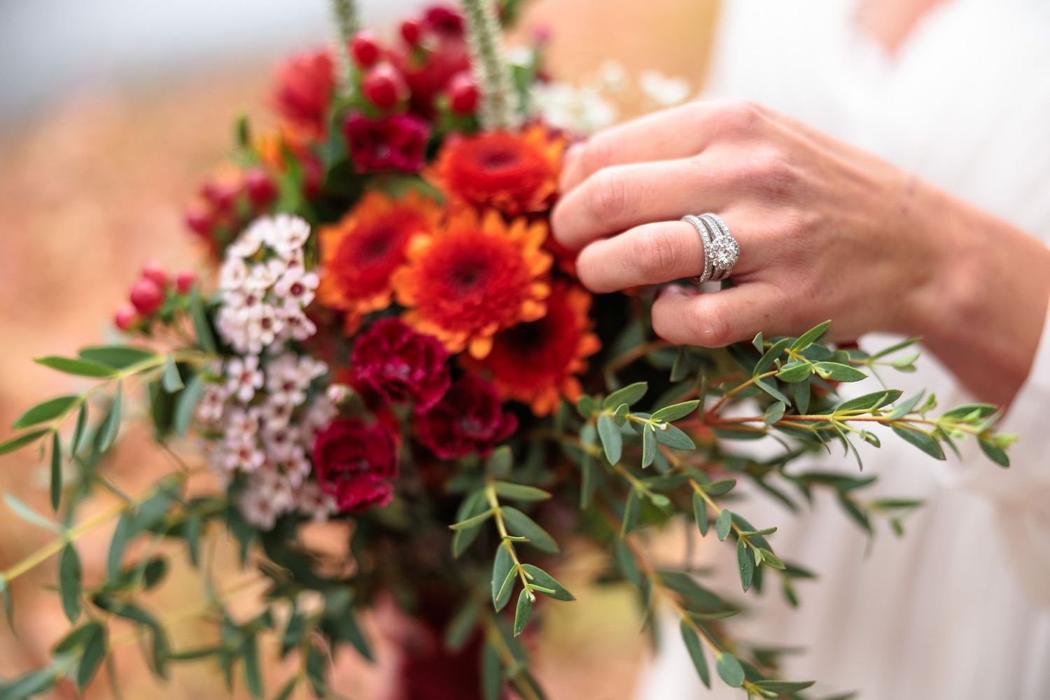 Close-up of bride's diamond ring beside vibrant red and orange wedding bouquet with greenery.