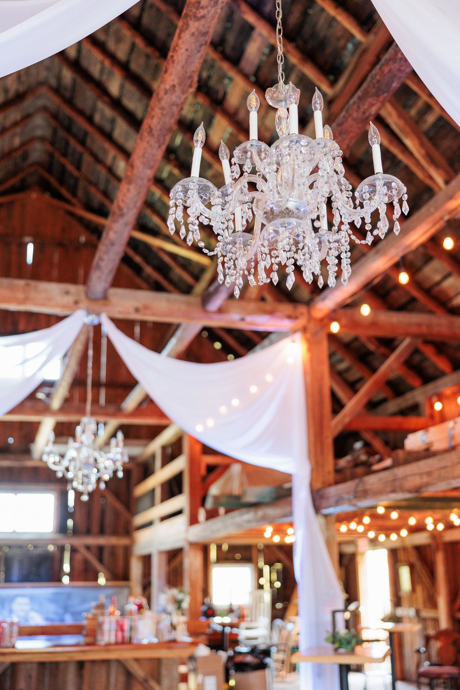 Crystal chandeliers and white draping decorate a rustic wooden barn ceiling.