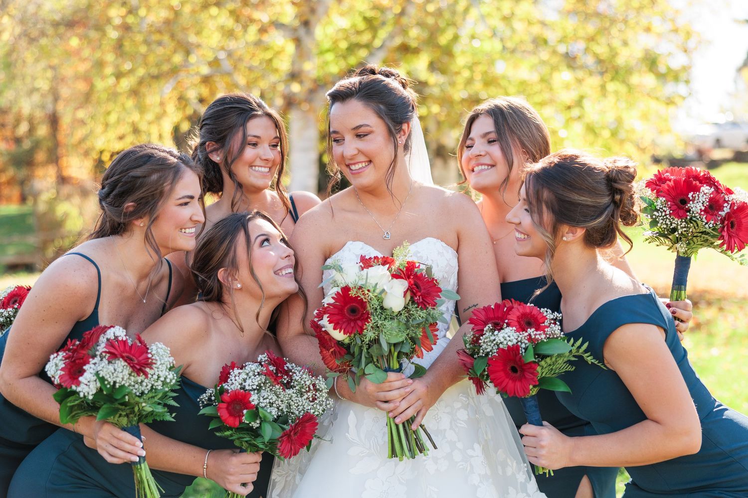 Bride in white lace gown laughs with five bridesmaids in teal dresses holding red floral bouquets outside.