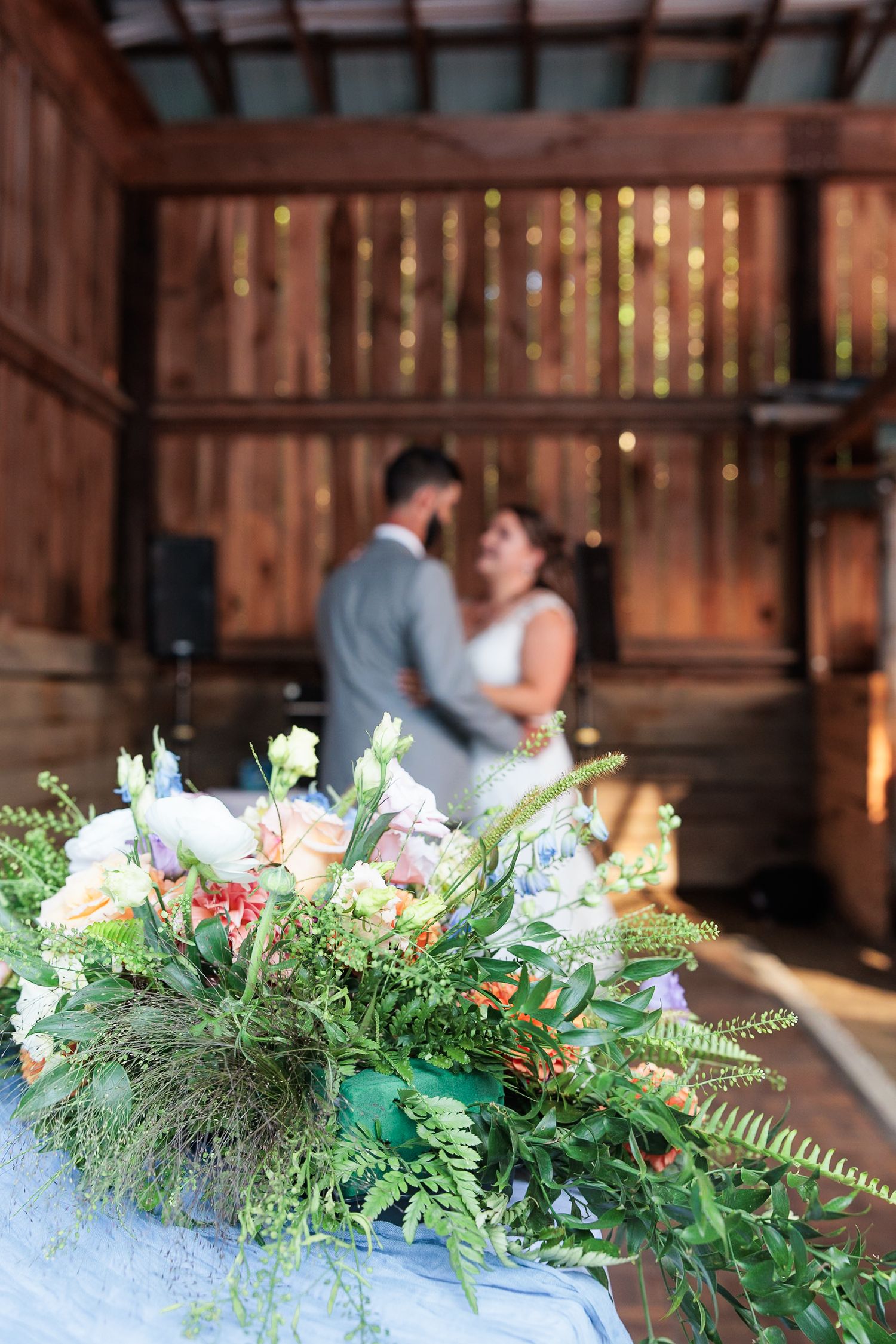 Colorful floral centerpiece in focus with bride and groom dancing softly blurred in barn background.