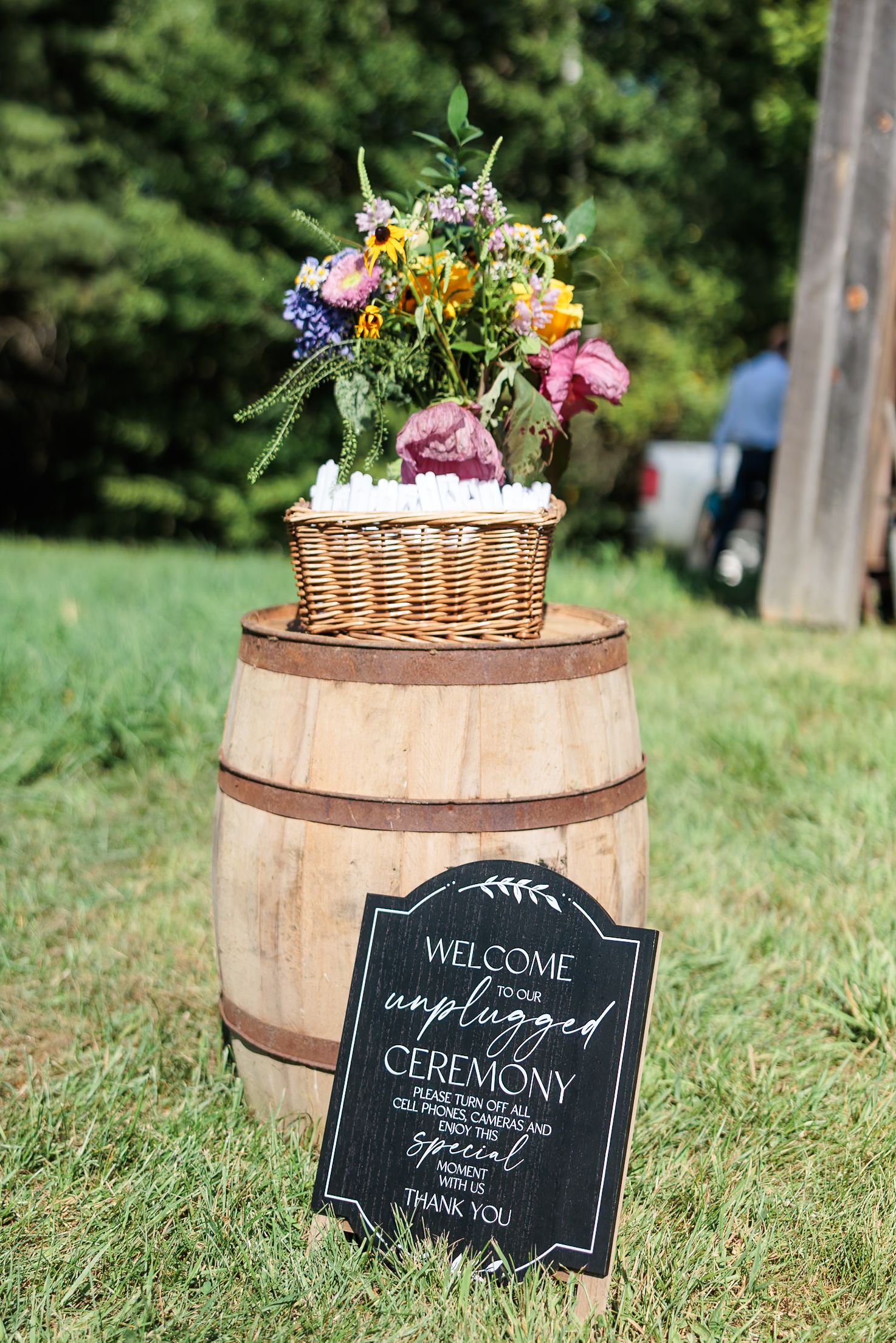 Rustic wooden barrel with welcome sign and floral basket greets guests at unplugged outdoor wedding ceremony.