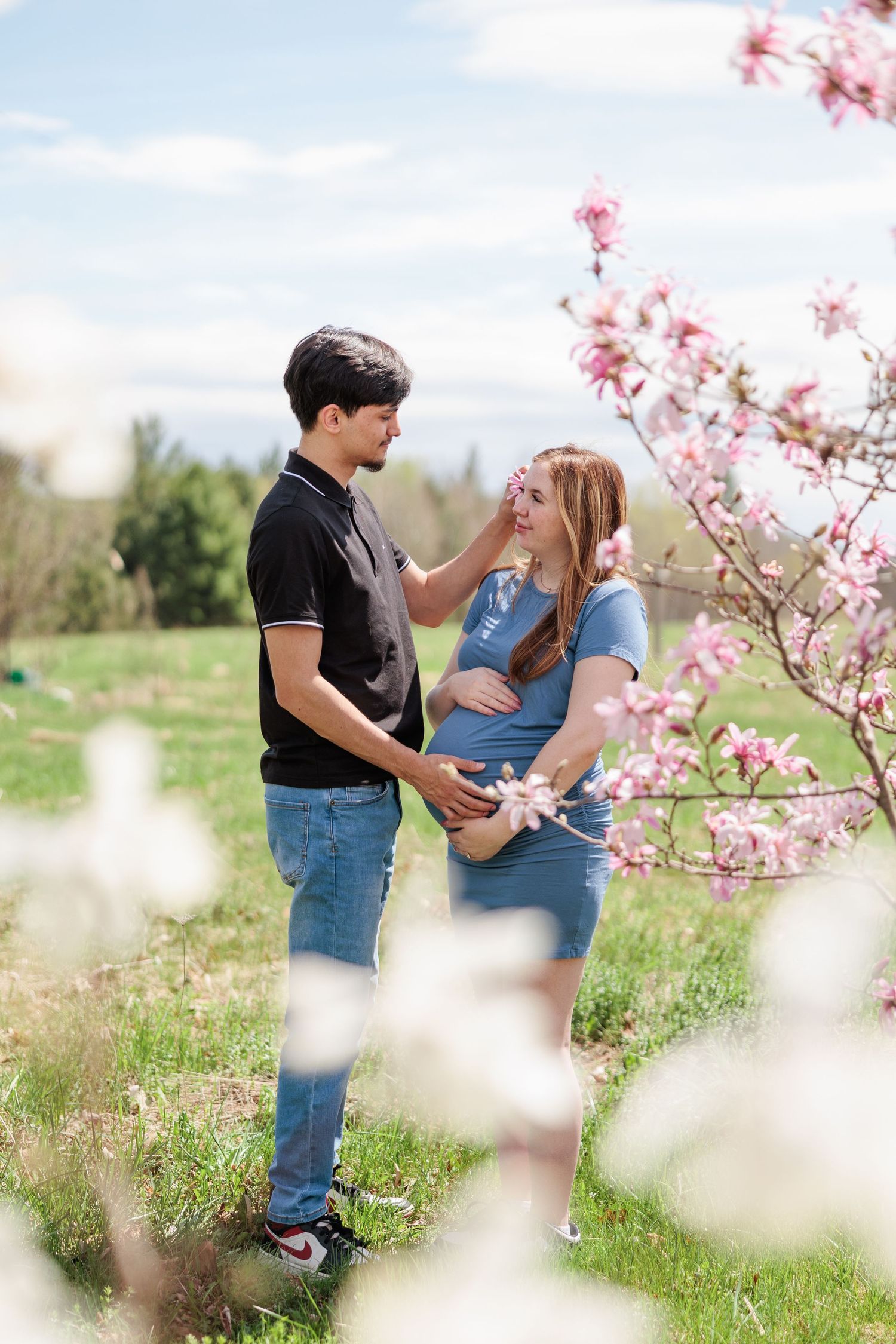 Expectant couple sharing a tender moment beside blooming pink magnolia trees on a sunny spring day.