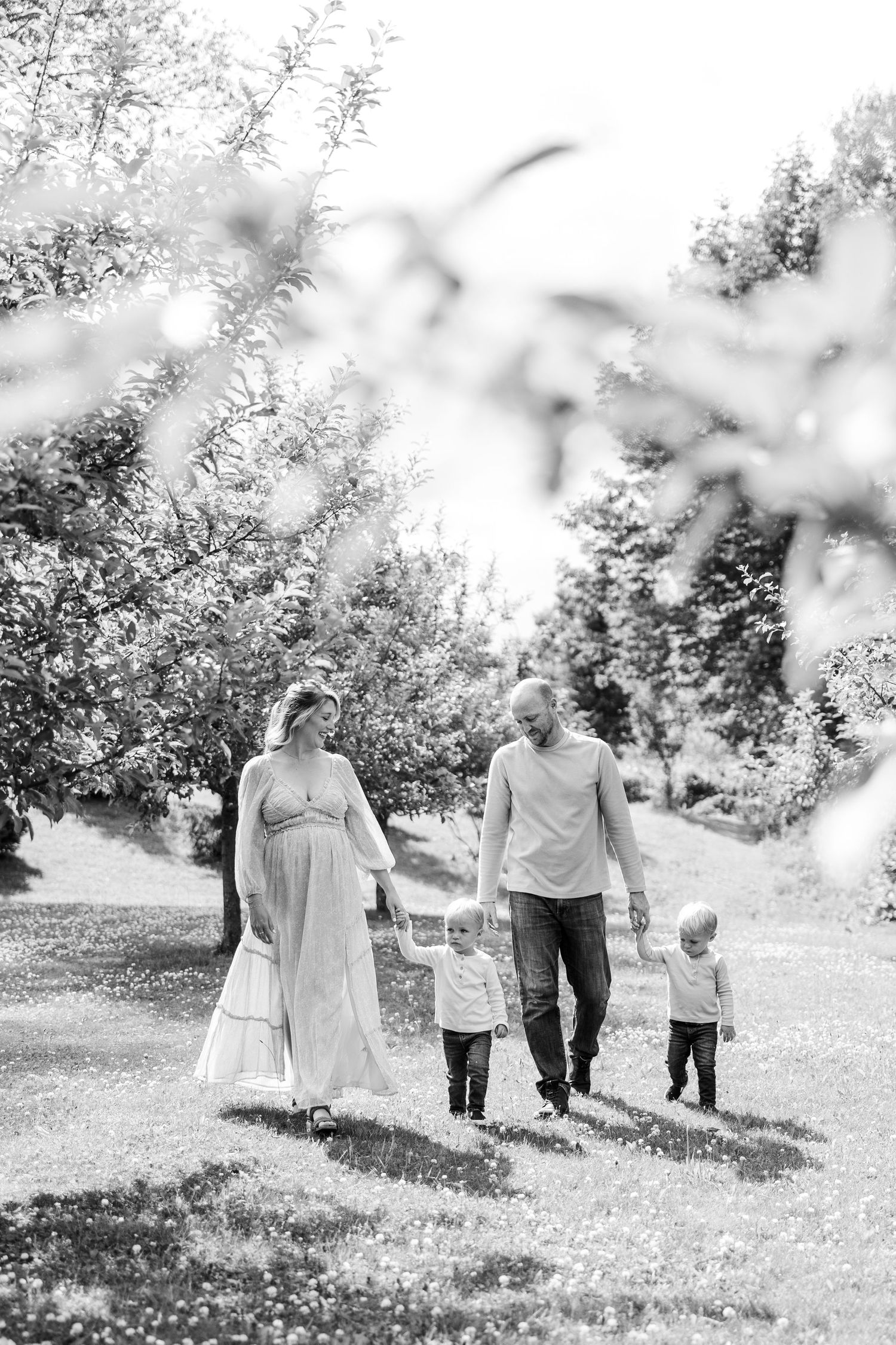 Black and white family photo of pregnant mother, father, and two children walking in orchard.