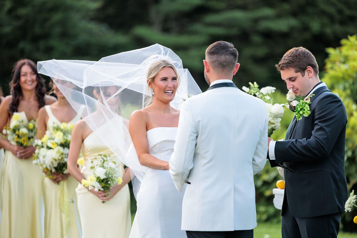Bride in white strapless gown exchanges vows with groom as bridesmaids in yellow dresses look on.