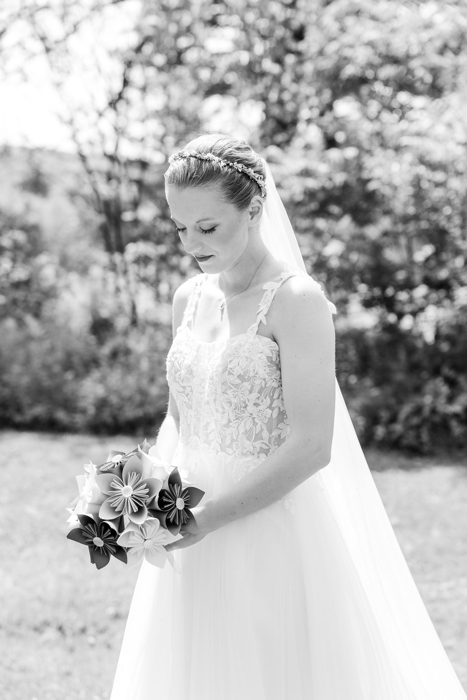 Black and white portrait of bride in lace gown holding origami flower bouquet outdoors.