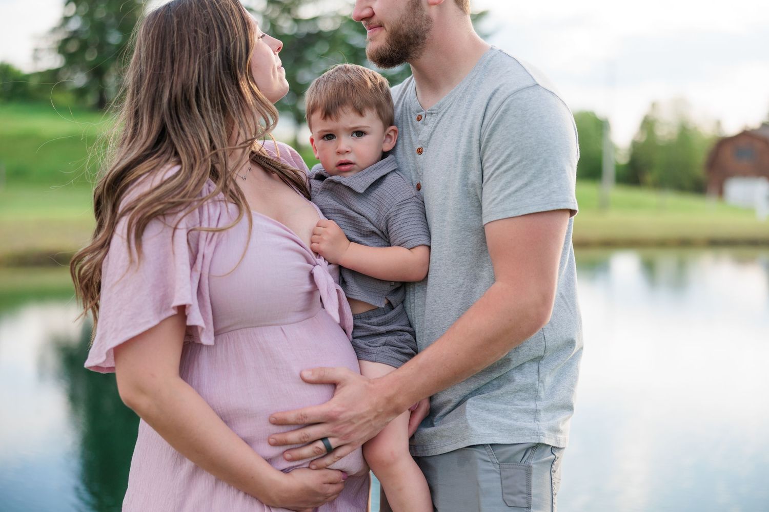 Couple holding toddler while man touches pregnant woman's belly by pond.