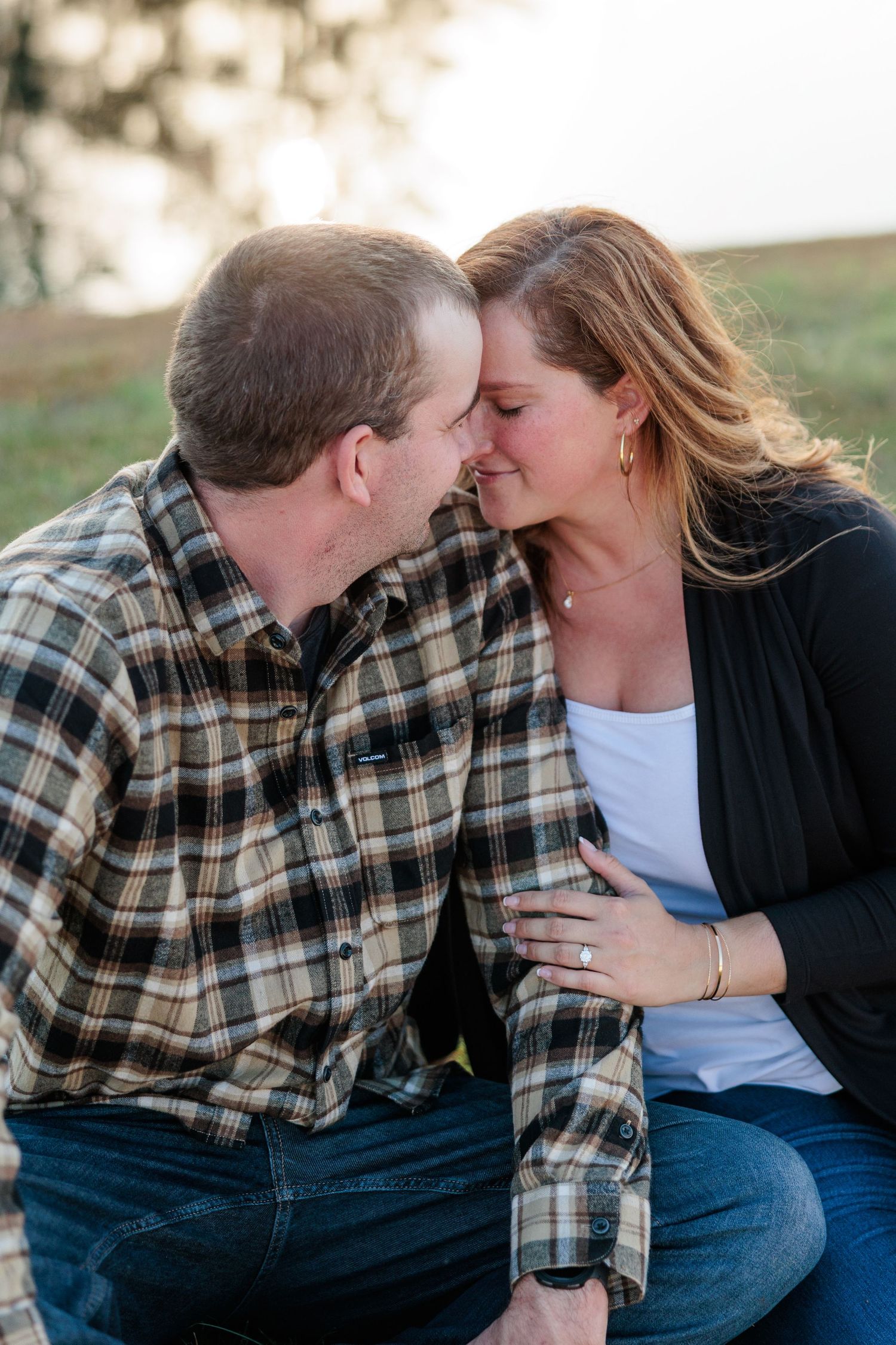 Engaged couple leaning in for a kiss while sitting together outdoors in warm autumn light.