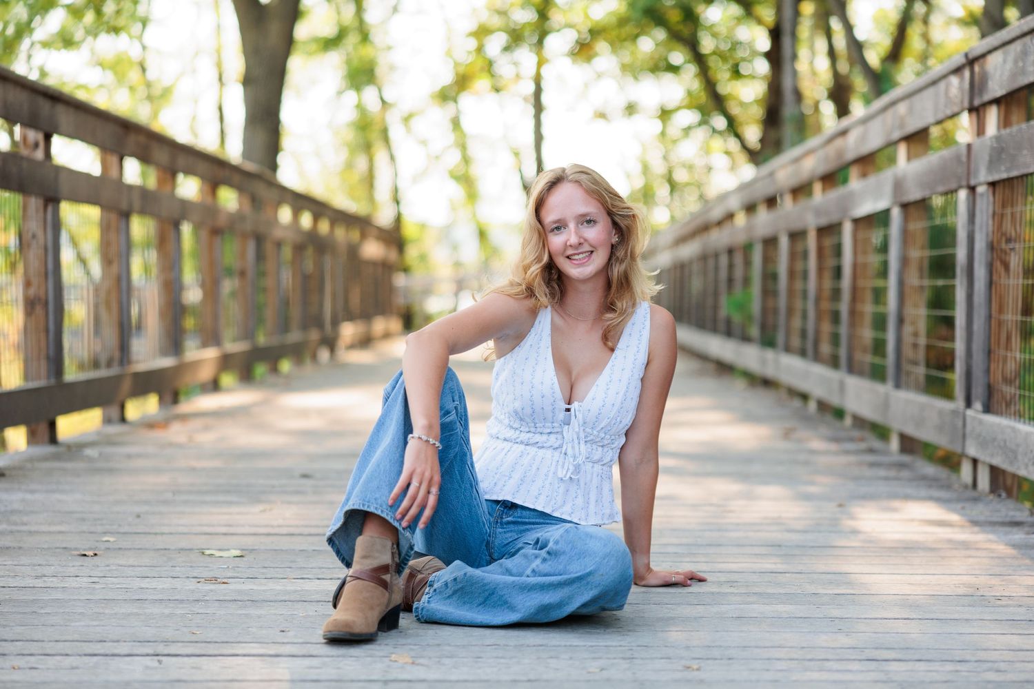 Young blonde woman smiling, sitting on wooden bridge in white top and jeans, surrounded by green trees.