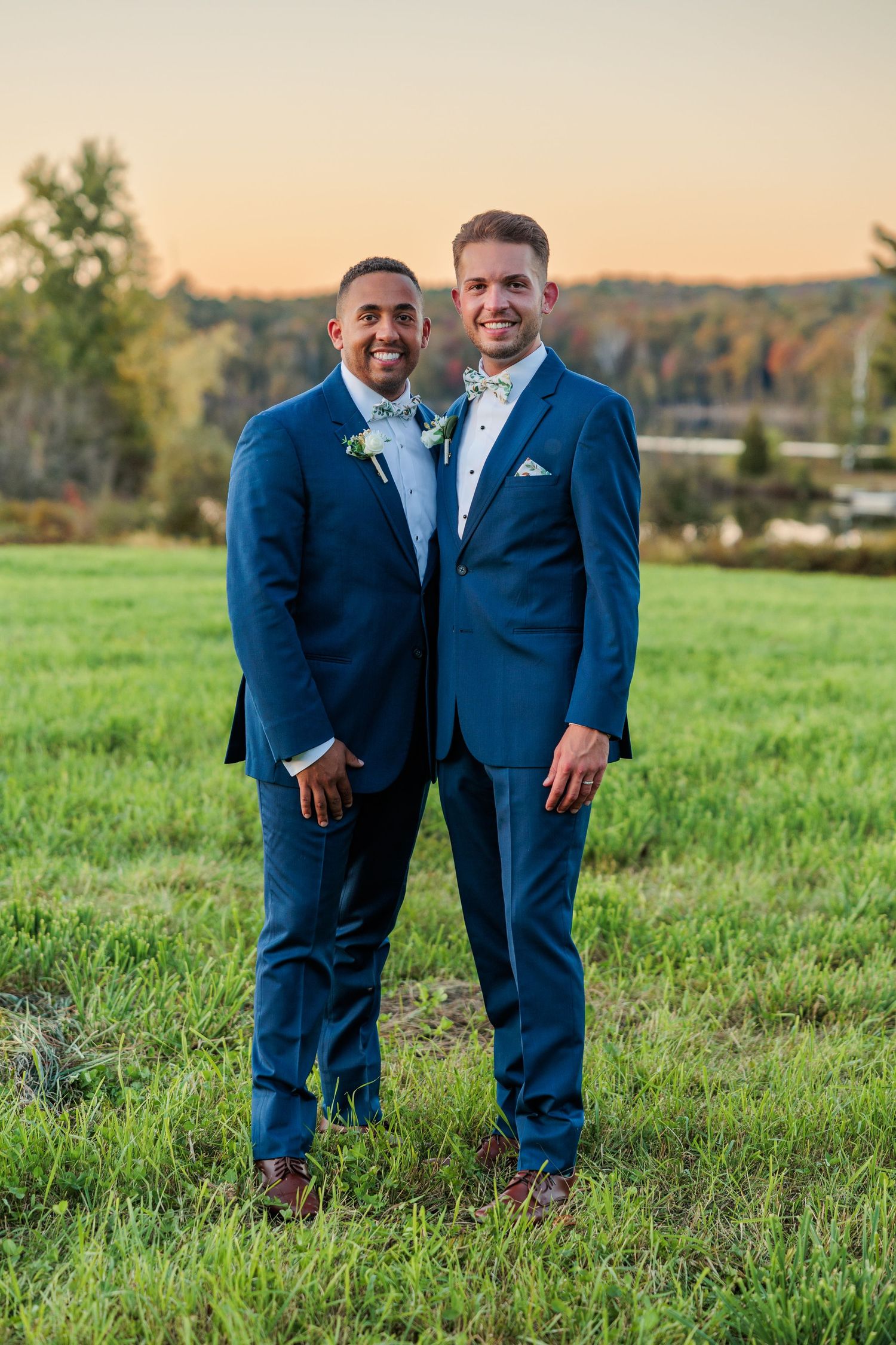 Two people in matching navy suits pose together in a green field at sunset.
