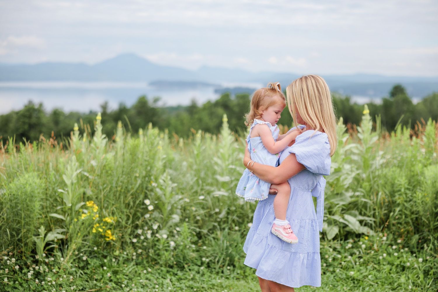A mother in a blue dress holds her young daughter in a matching outfit in a lush green field with mountains behind.