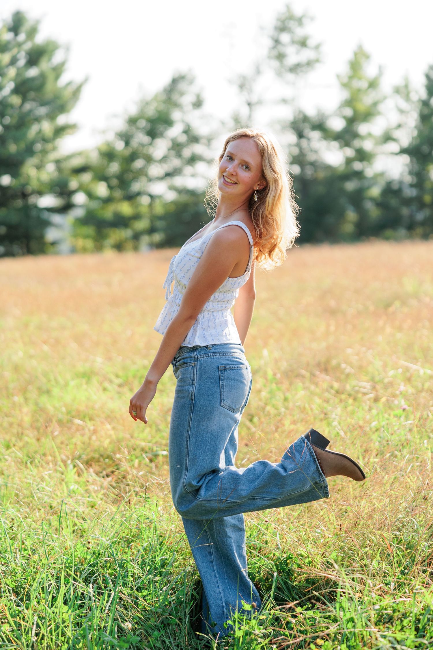 Young woman in white top and jeans smiling in a sunny field with green trees in the background.