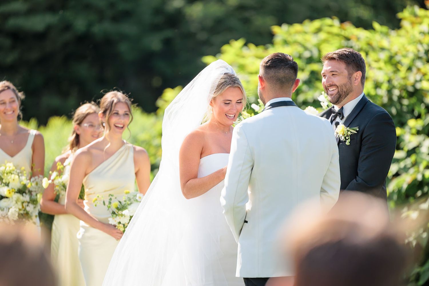 Bride in white ball gown laughs with groom in white tuxedo jacket during outdoor wedding ceremony.