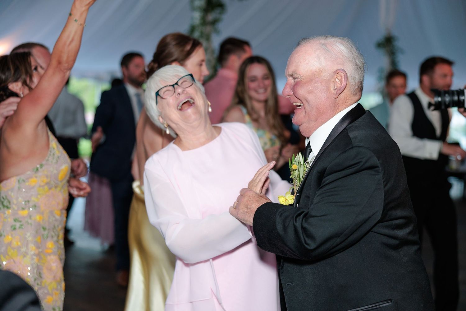 Elderly couple laughing and dancing joyfully at an outdoor wedding reception under a tent.