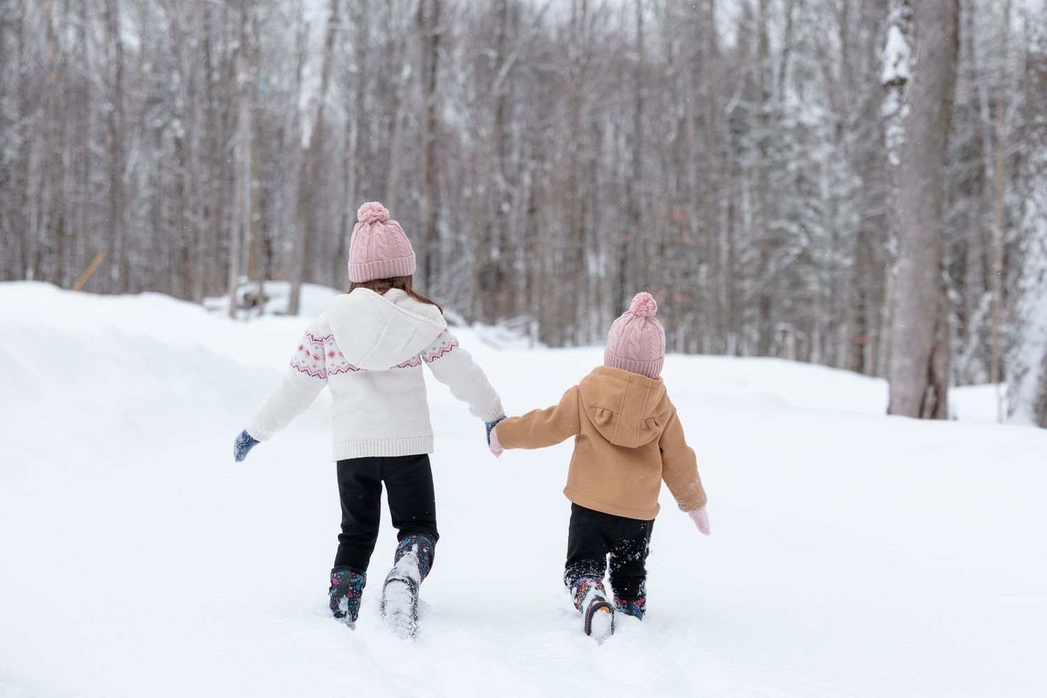 Two young girls in pink winter hats hold hands walking through a snowy forest.