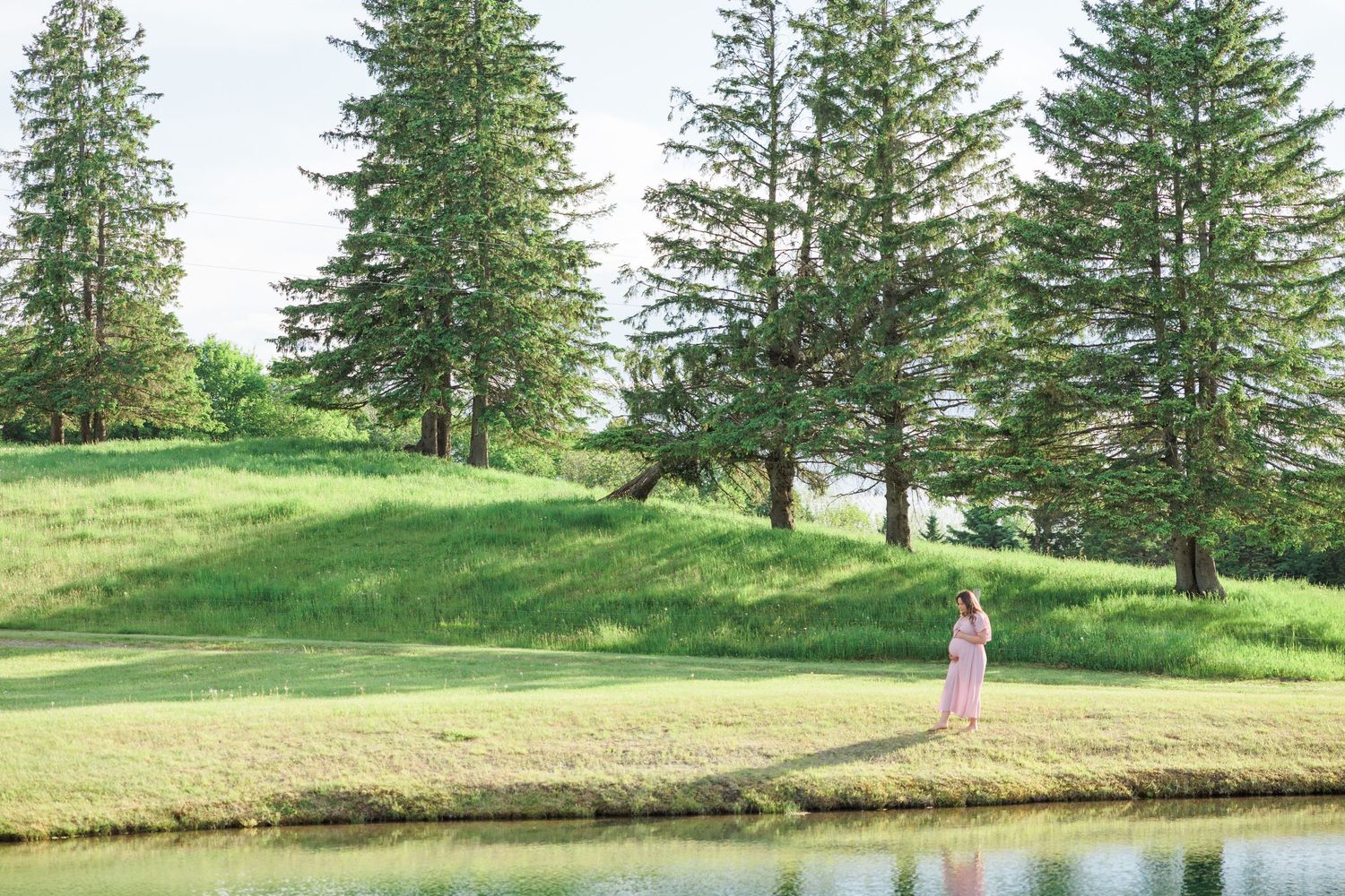 Pregnant woman in pink dress standing alone near a pond surrounded by tall evergreen trees on a green hillside.