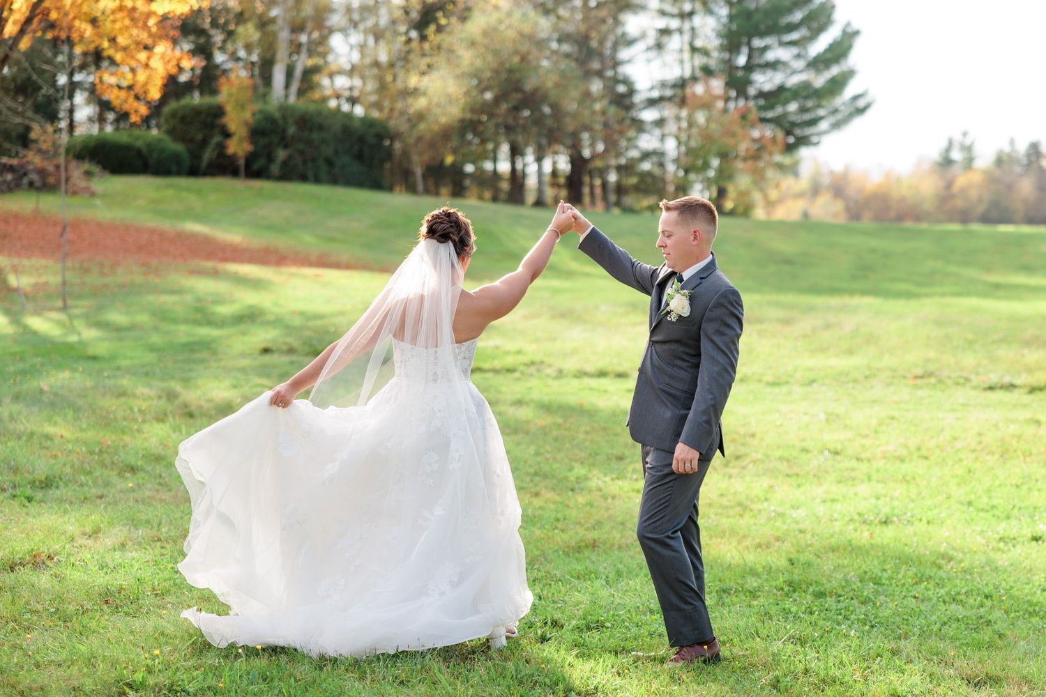 Groom spinning bride in flowing white gown on green lawn with autumn trees behind.