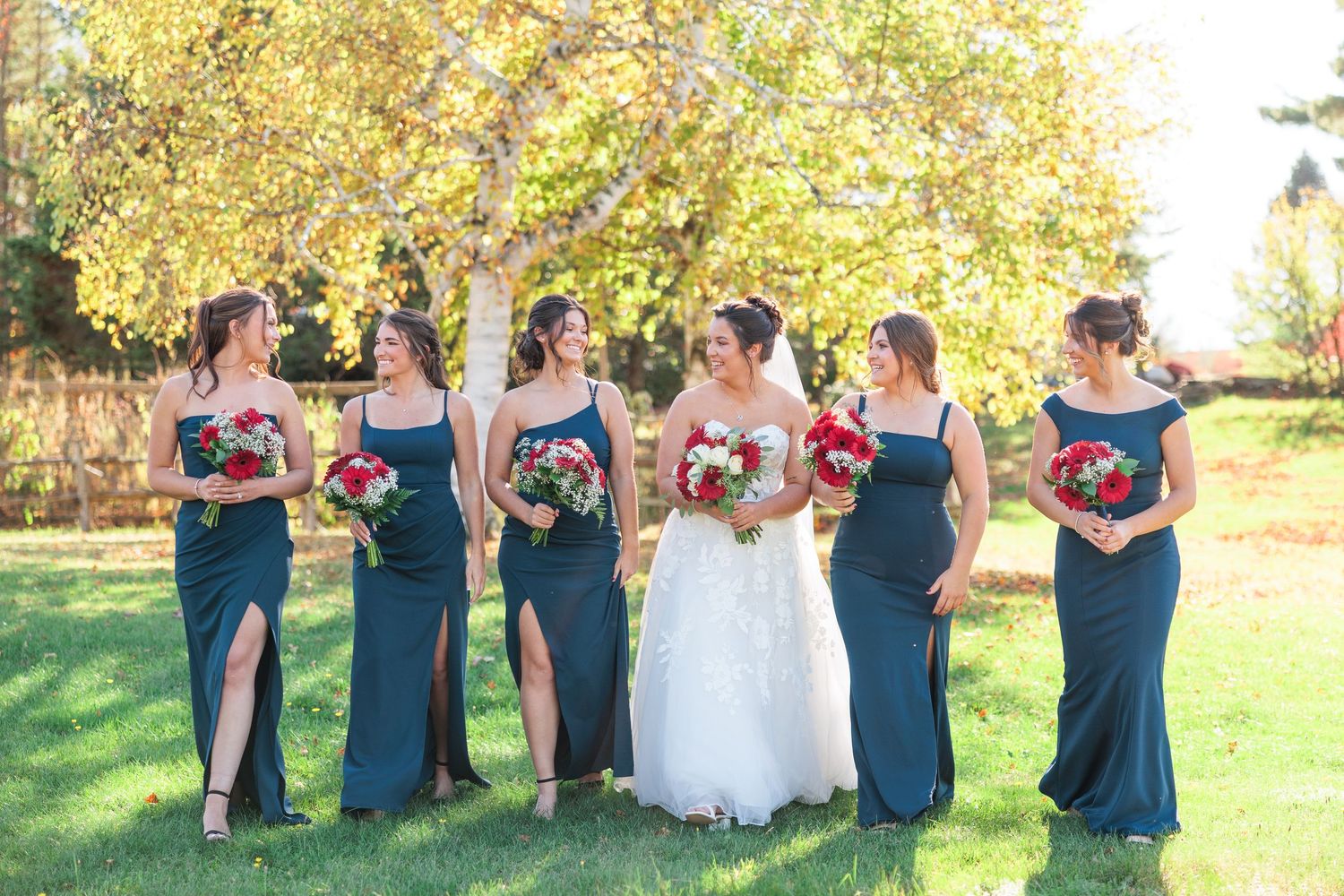Bride in white gown walks with five bridesmaids in teal dresses carrying red bouquets under autumn trees.