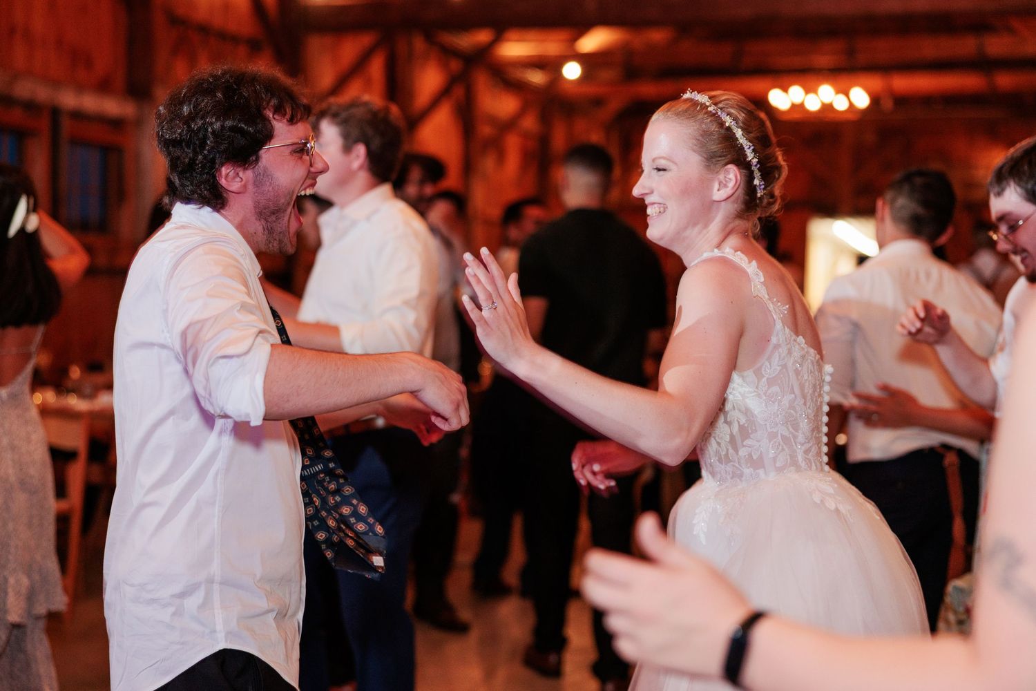 Bride in lace gown laughs and dances with a curly-haired guest at a lively barn wedding reception.