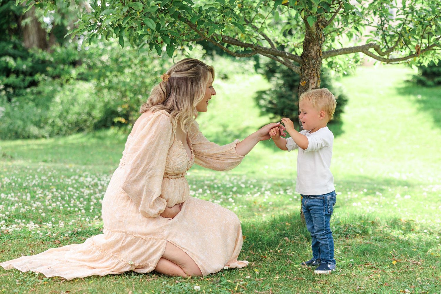 Pregnant woman in peach dress kneeling on grass as toddler hands her something outdoors.