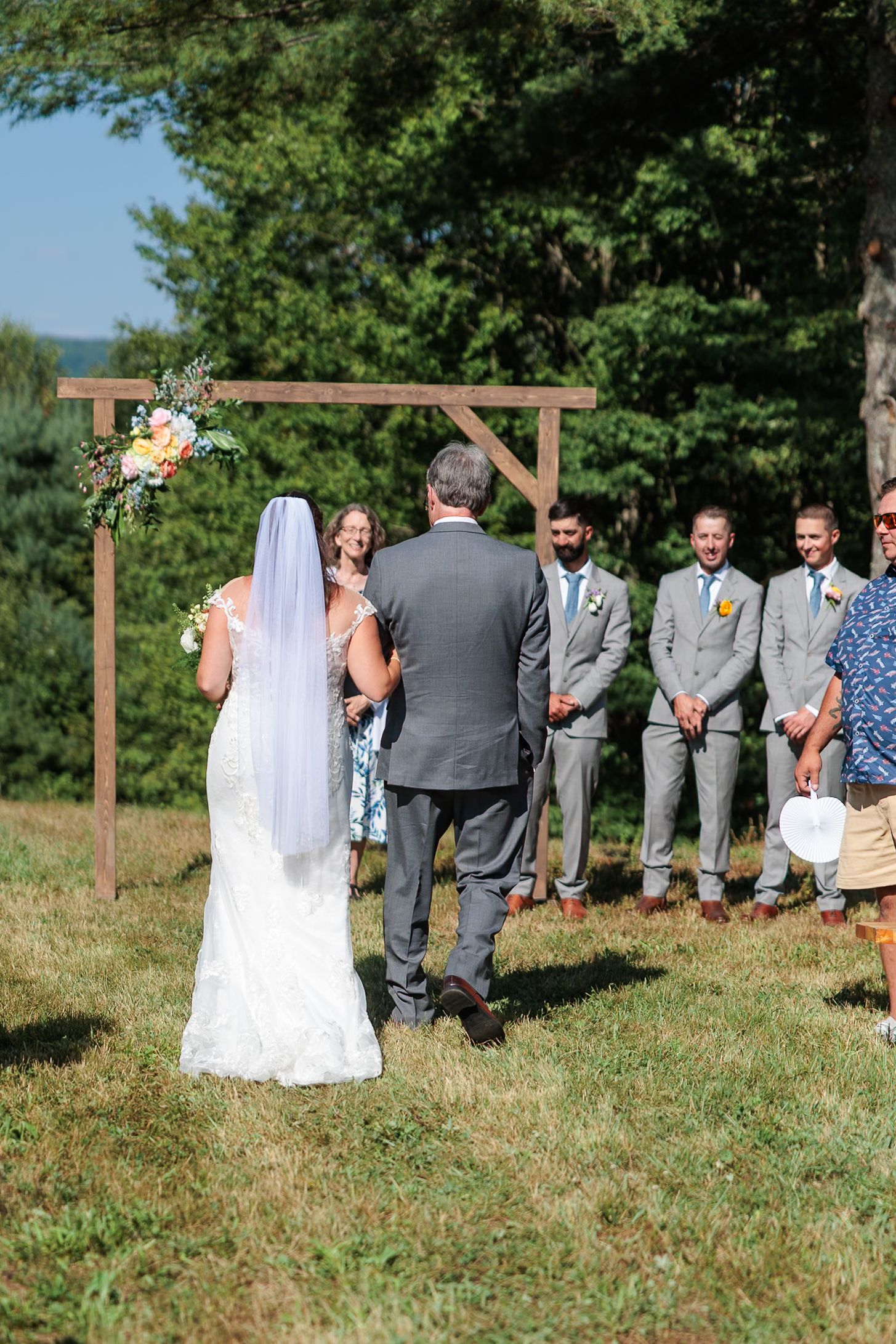 Bride and groom stand at rustic wooden arch altar with groomsmen lined up during outdoor wedding ceremony.