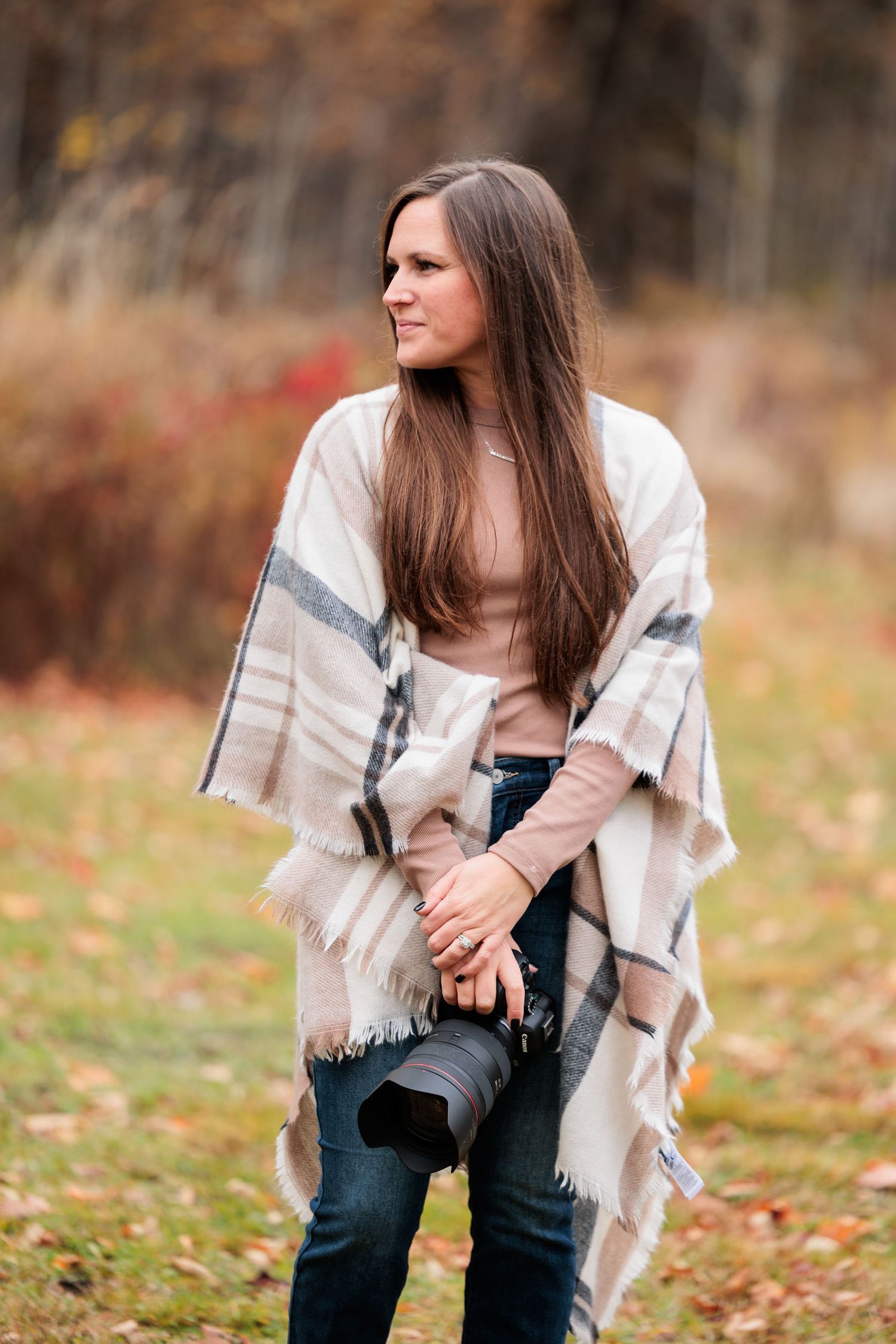 Woman in beige plaid poncho holding a camera standing in a fall foliage outdoor setting.