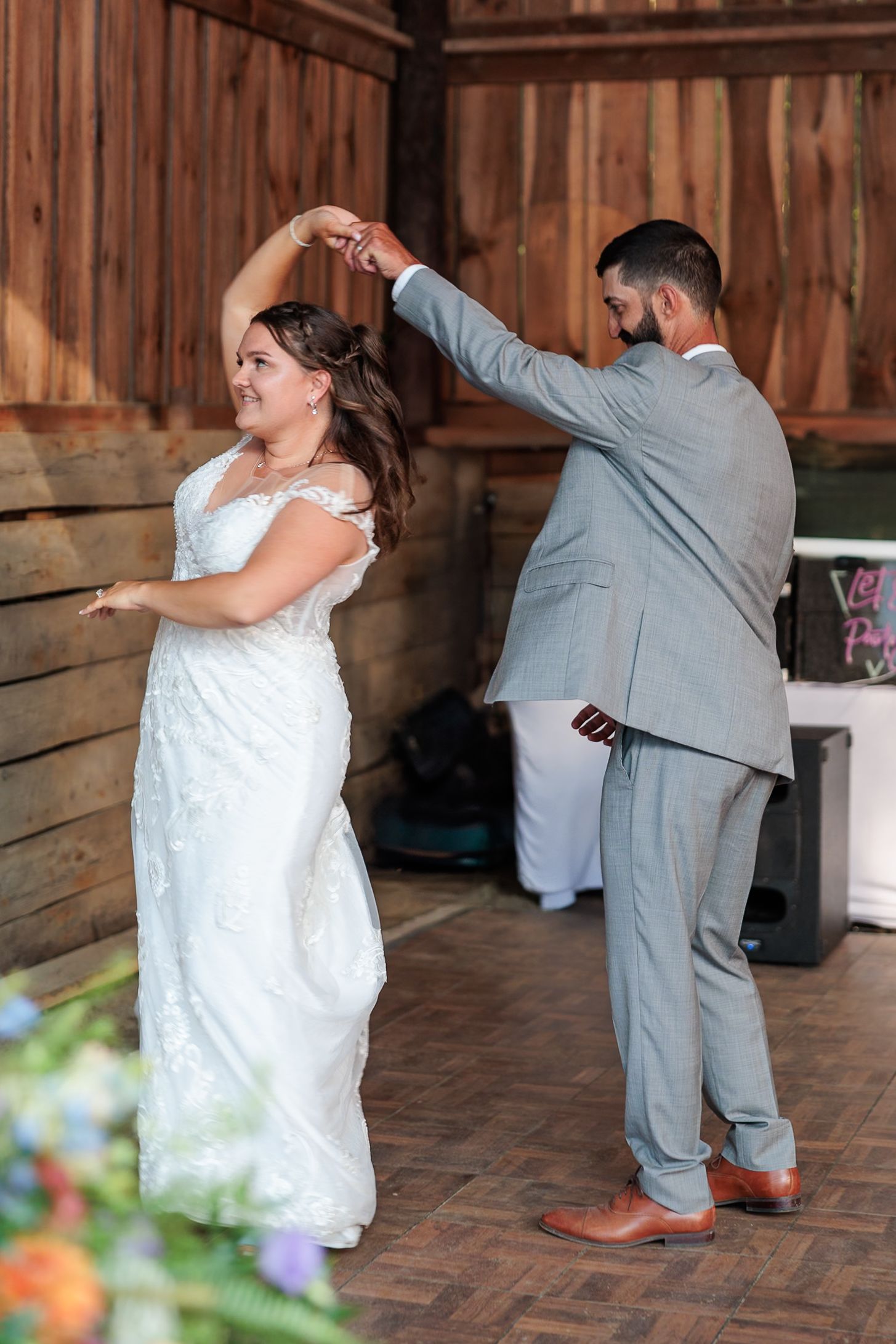Groom spinning bride in white lace dress during first dance inside a rustic barn venue.
