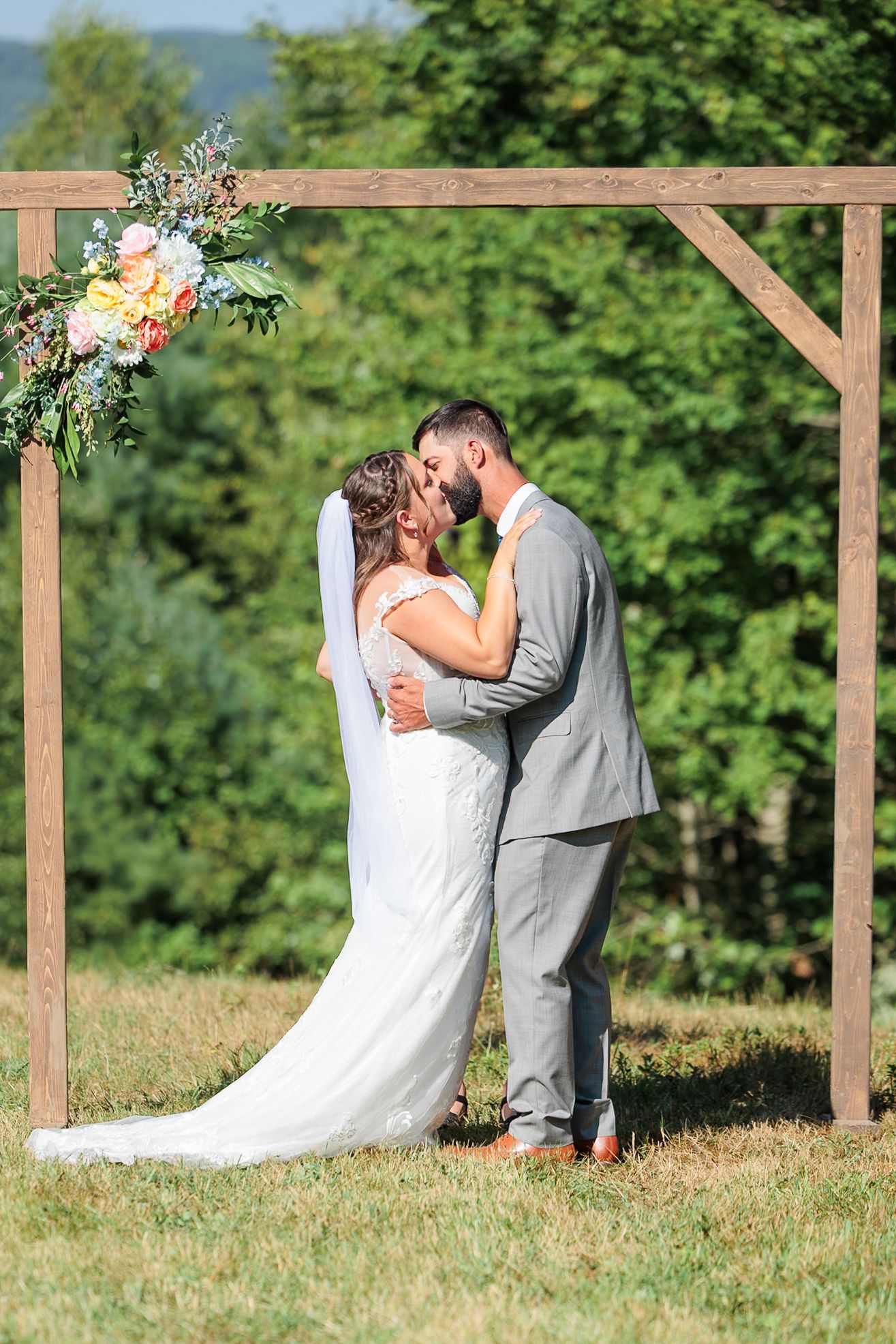 Bride and groom share first kiss under rustic wooden arch adorned with colorful flowers at outdoor wedding ceremony.
