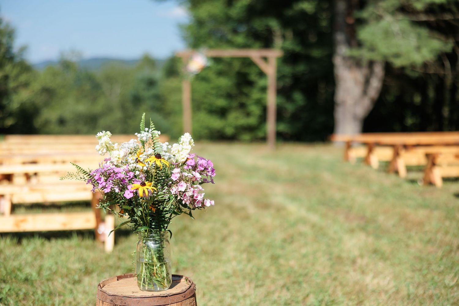 Mason jar with wildflowers on barrel decorates aisle of outdoor wedding ceremony with wooden benches.