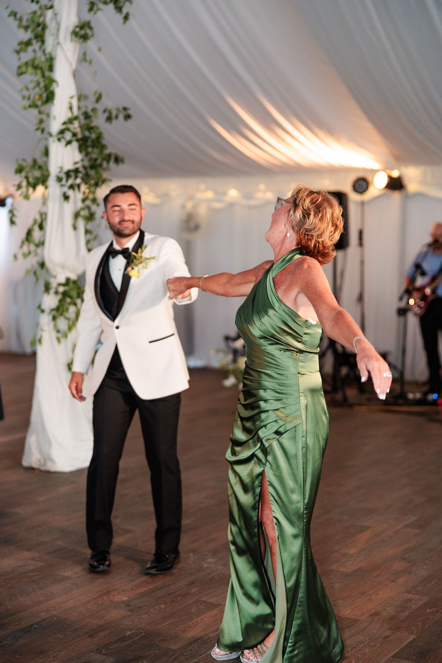 Groom in white tuxedo jacket dances with mother in green gown at wedding reception.