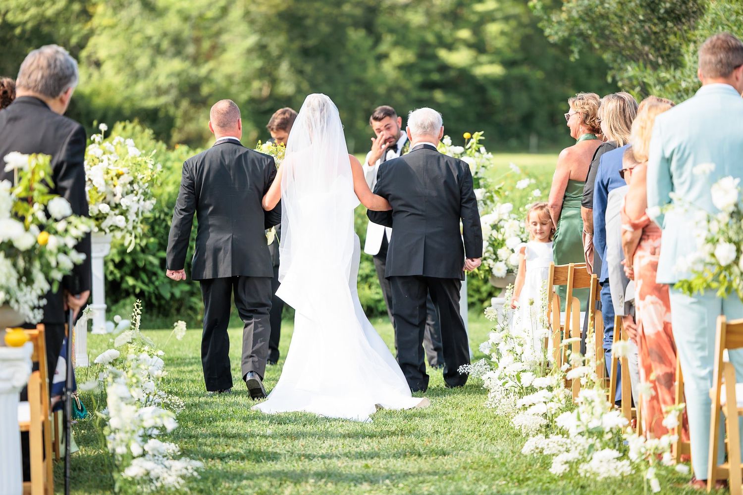 Bride walks down outdoor aisle in white gown and veil with escorts at garden wedding ceremony.