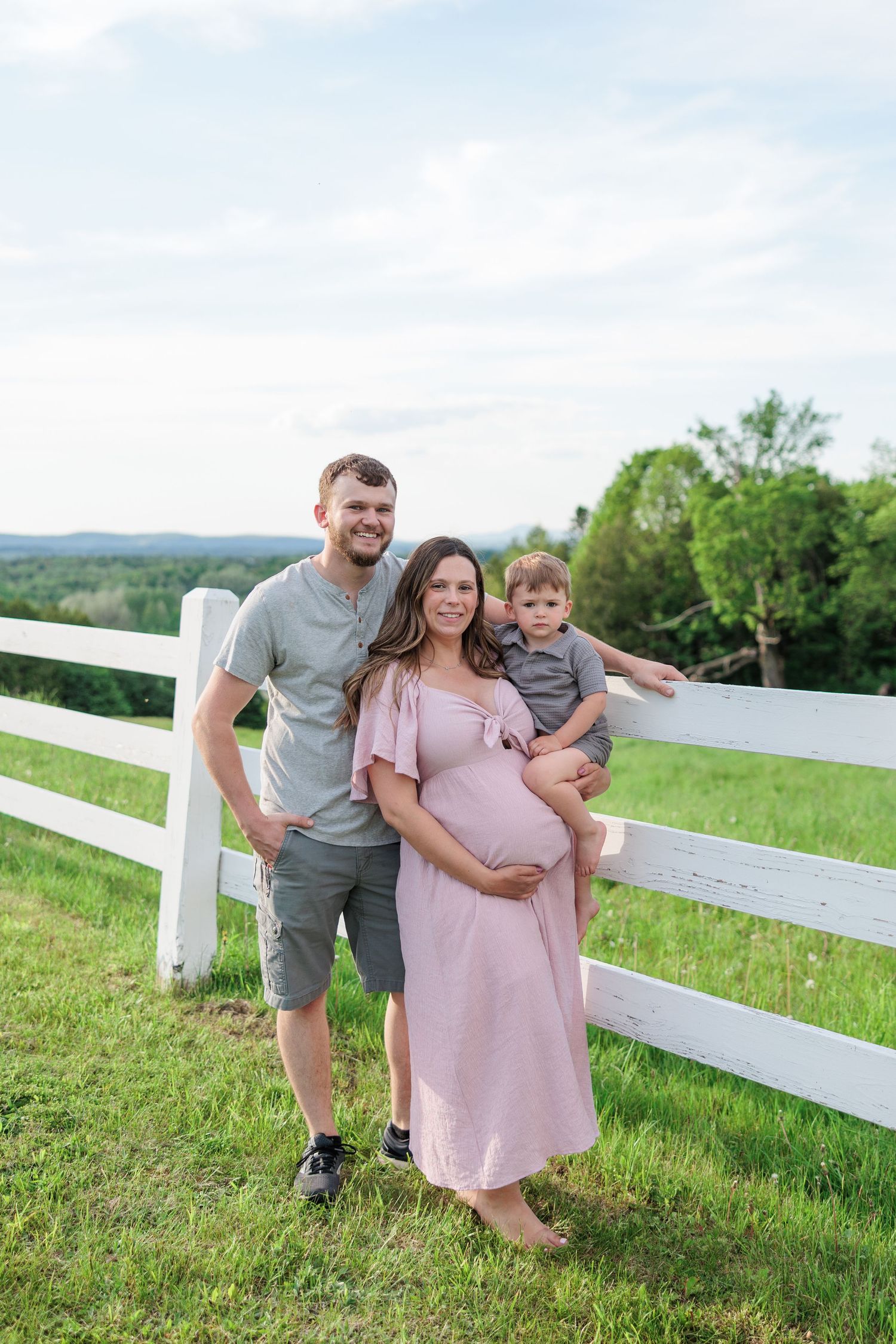 Family of three with pregnant mom poses by white fence on green hillside.