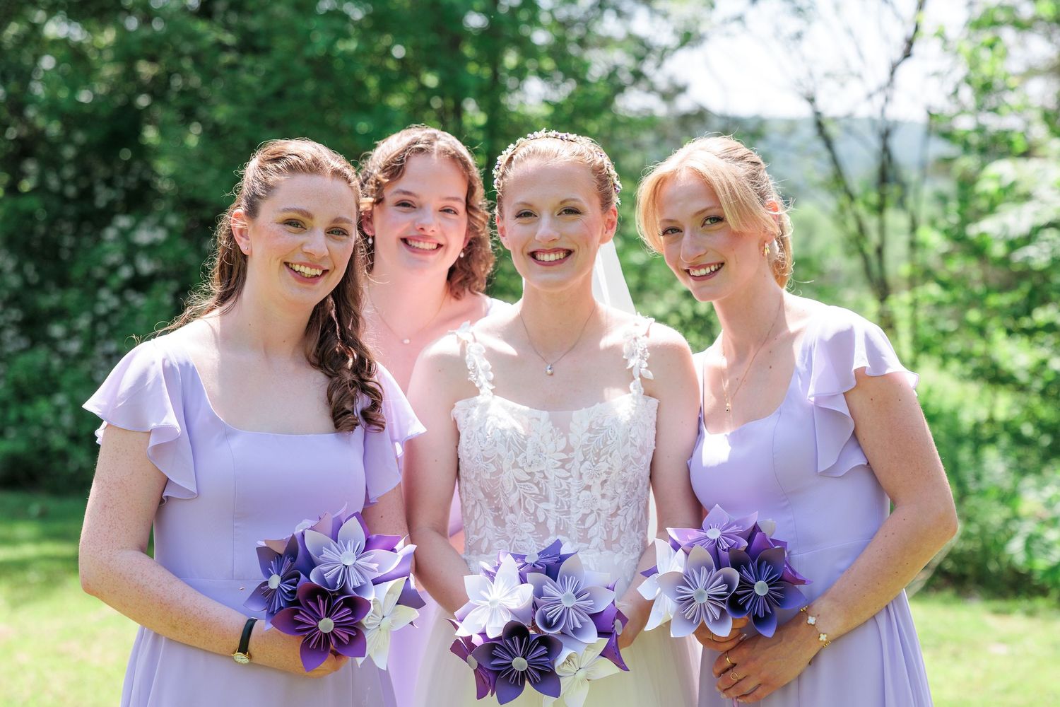 Bride with three bridesmaids in lavender dresses holding purple origami bouquets outdoors.