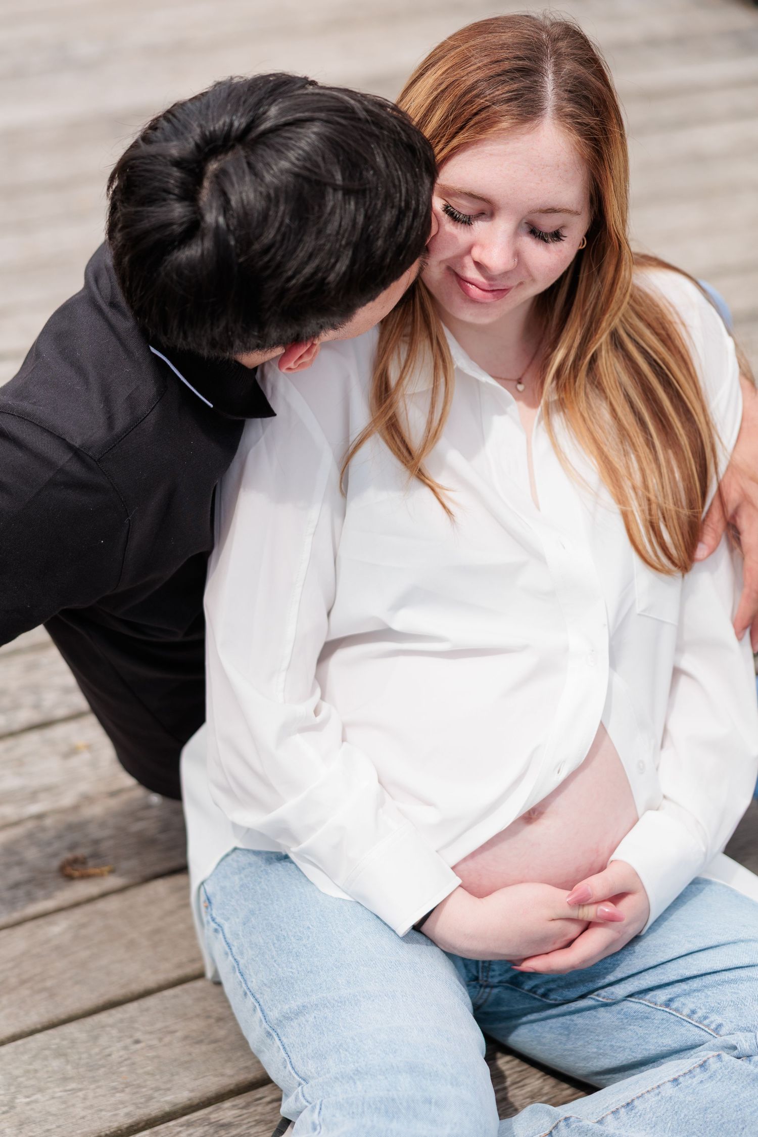 Couple sharing tender moment on wooden deck, man kissing pregnant woman's cheek as she cradles her baby bump.