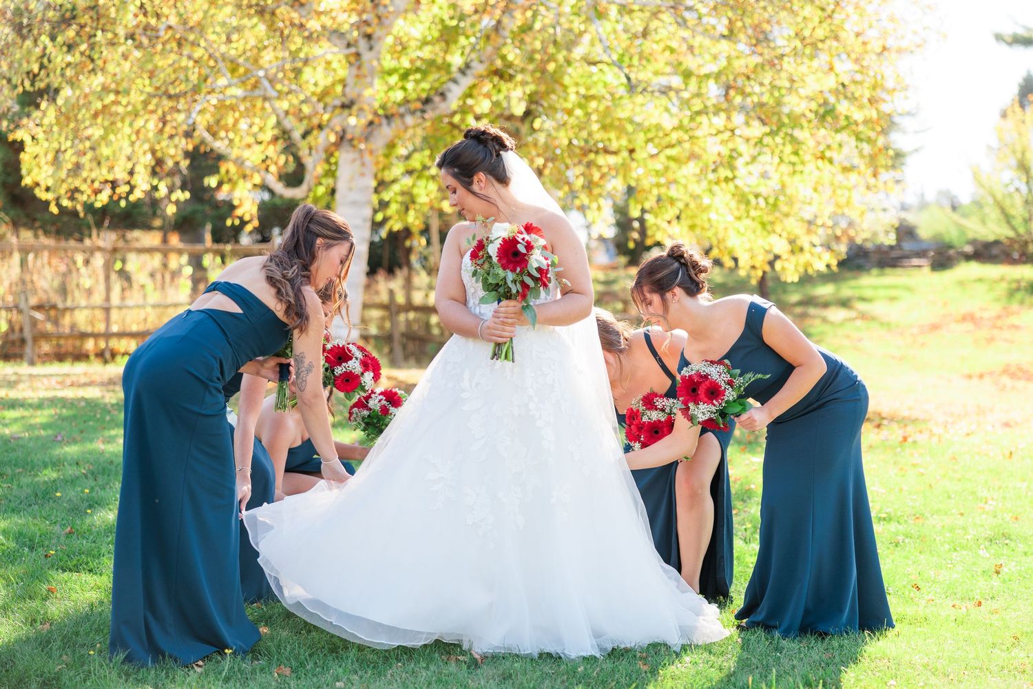 Bridesmaids in teal gowns help adjust bride's white ball gown dress outdoors under a yellow autumn tree.