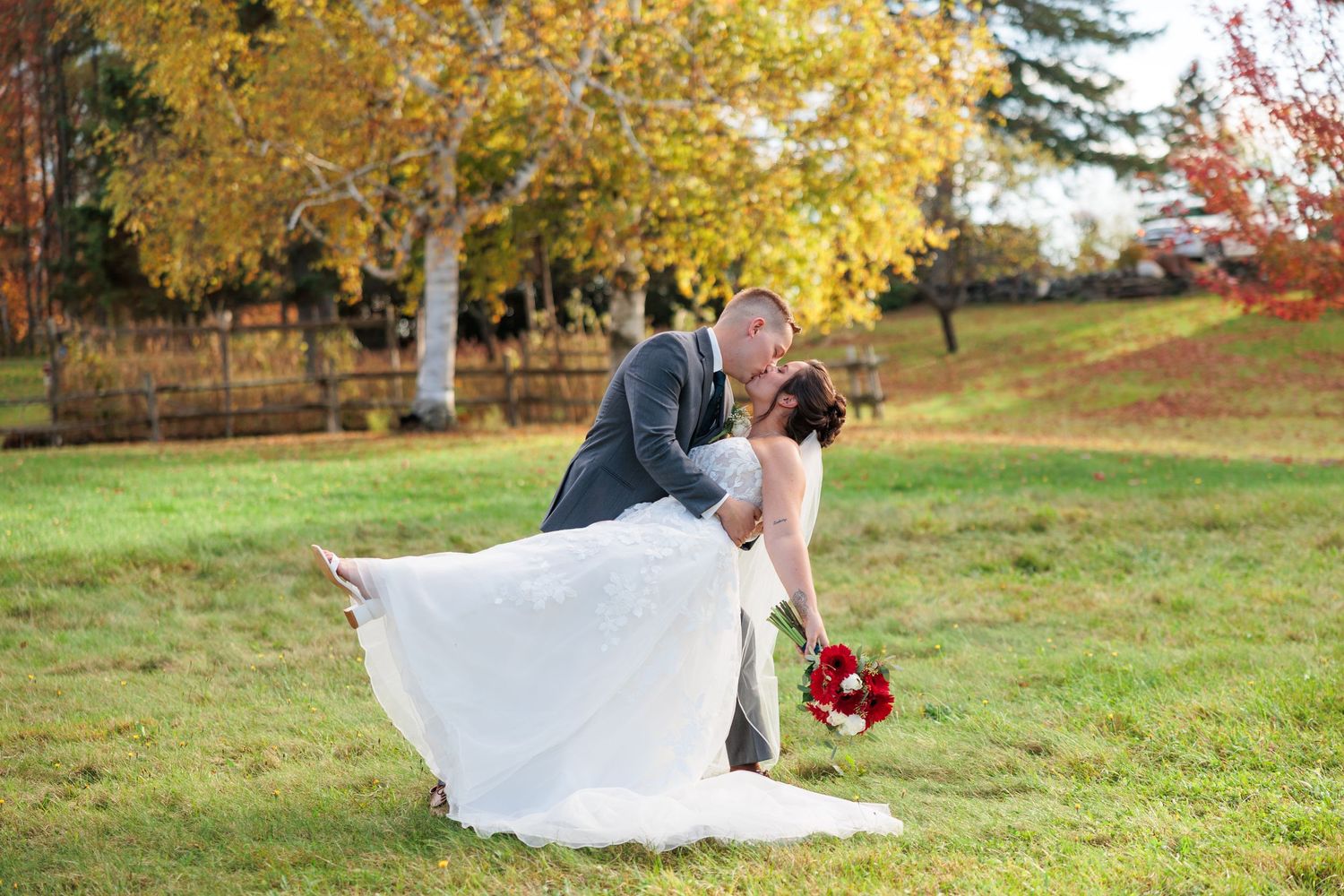 Groom dipping bride dramatically on green lawn surrounded by colorful autumn foliage.