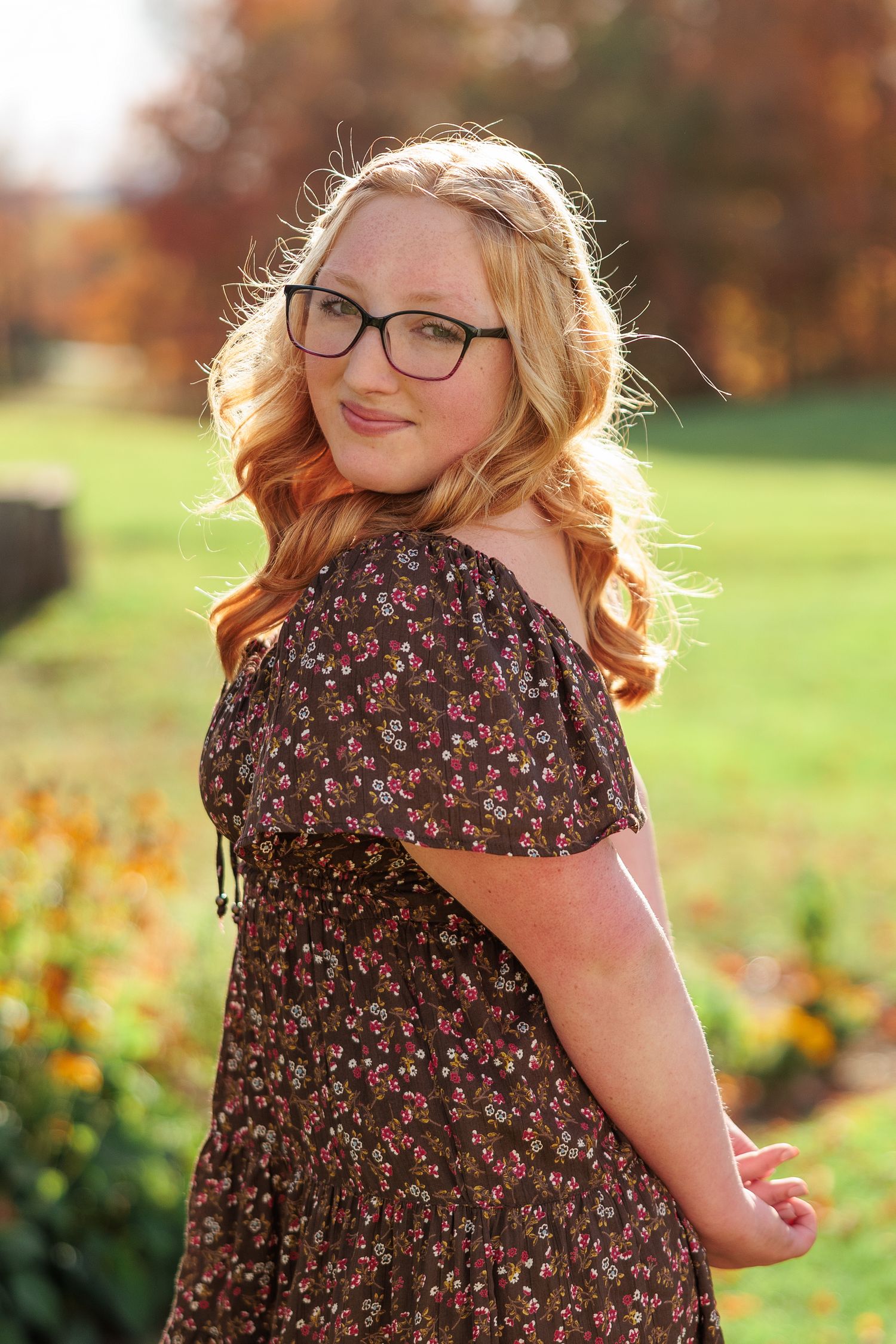 Young blonde woman in floral dress and glasses smiling outdoors in autumn park with colorful fall foliage.